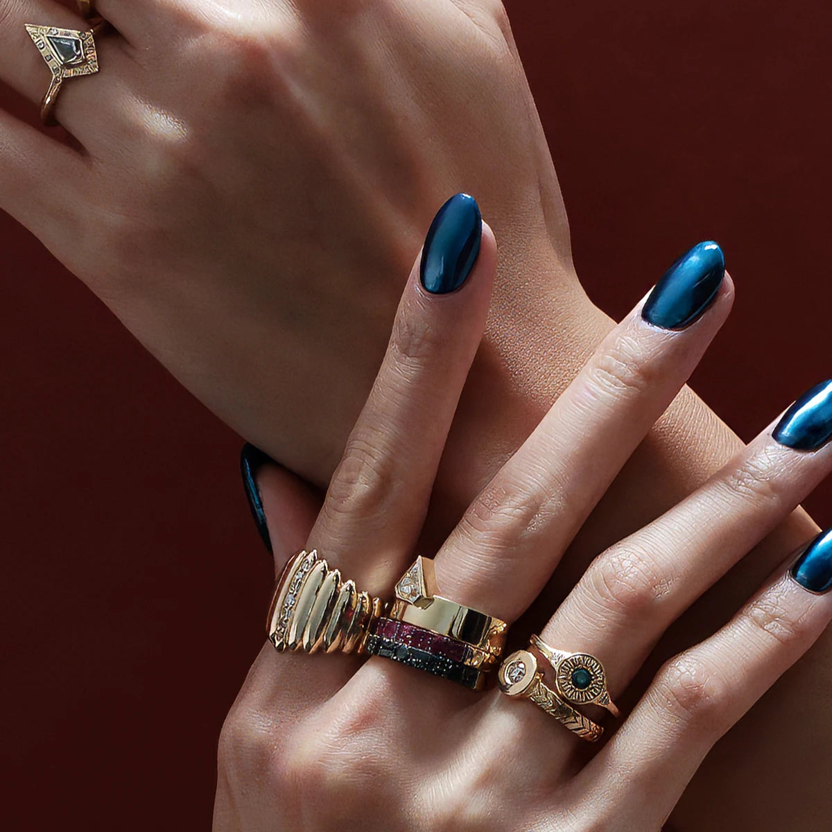 Two hands in close shot studio photography are modelling for fine jewelry pieces. The hands have dark blue metallic nails. The bakcground is red wine color. The hands wear 7 unique designer rings of unique different styles. All of them are in yellow gold. Gemstones seen are salt & pepper diamond, raw garnet, white sapphire, natural diamond, rubies and black diamonds.