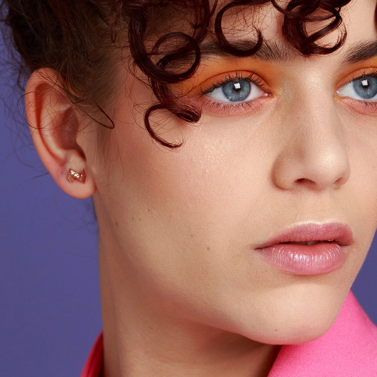 Young lady&#39;s face with blue eyes and curly hair photographed very close up. She wears fluorescent pink clothing and is photographed in the studio against a purple background. She is wearing a pink earring with diamonds. The earring is shaped like a lightning bolt, and is a creation of the Canadian jewellery brand Bena Jewelry. 