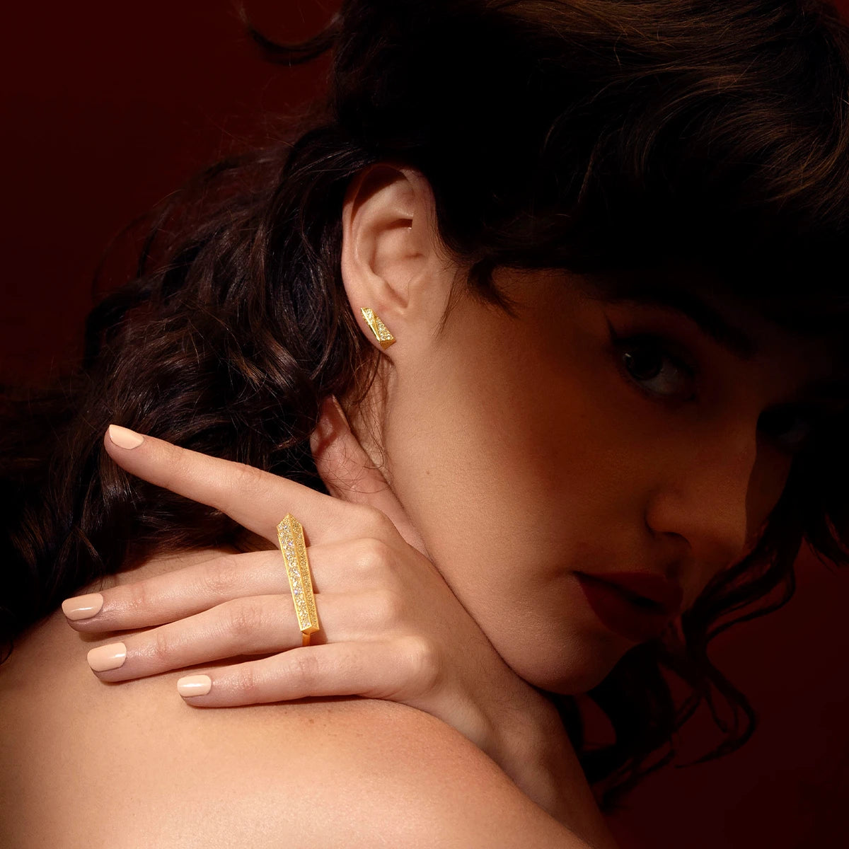 Fashion photography of a young woman with curly brown hair whose face is in shadow. She looks at the camera and raises her hand over her shoulder: on her hand, illuminated by the light, she wears a unique high end ring that looks like an objet d&#39;art. It&#39;s a large yellow gold baguette set with several diamonds. She is also wearing a matching diamond earring. The jewels have a modern, geometric look, created by independent Canadian designer Bena Jewelry.