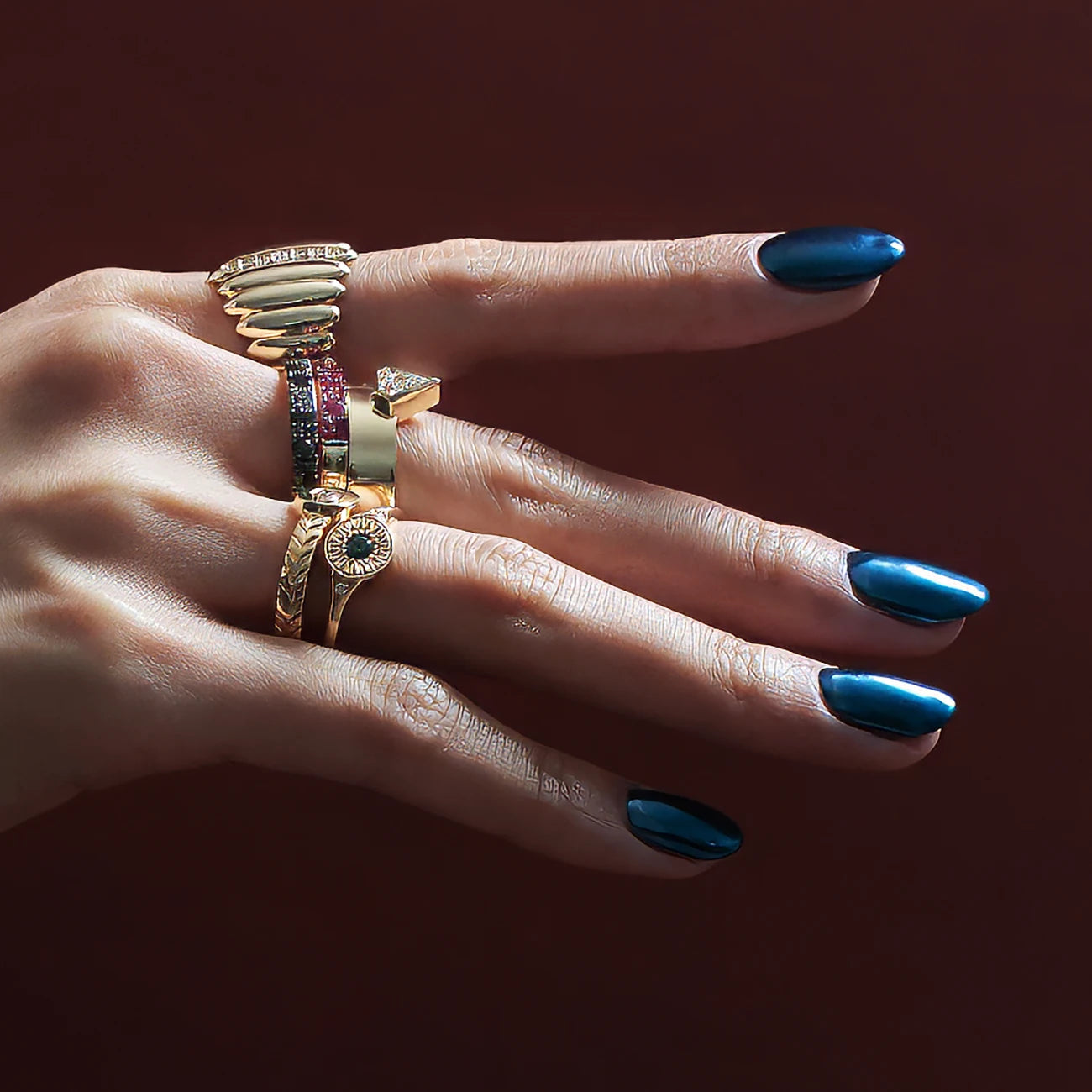 A hand in close shot studio photography is modelling for fine jewelry pieces. The hand has dark blue metallic nails. The background is red wine color. The hand wears 6 unique designer rings of unique different styles. All of them are in yellow gold. Gemstones seen are diamonds, white sapphire, green sapphire, natural black diamonds and rubies.