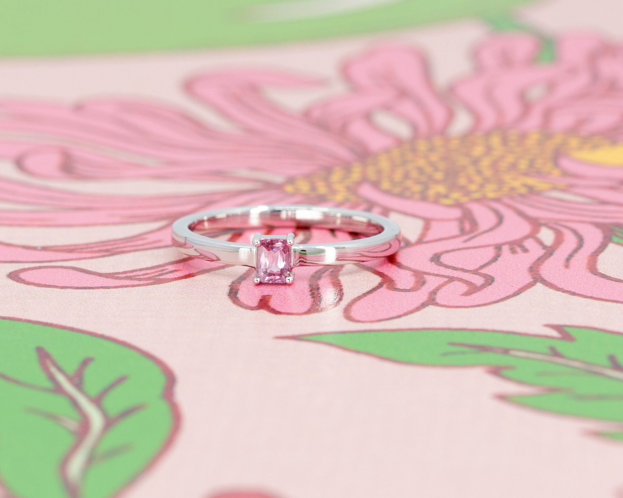 Elegant white gold ring with a pink sapphire baguette, displayed on a soft pink background. Designed by Bena Jewelry in Montreal, Canada.