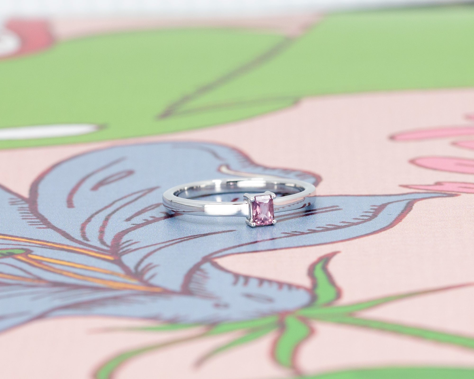 Boxy white gold ring with a pink baguette sapphire, showcased on a pastel pink background. A bold design by Bena Jewelry in Montreal.