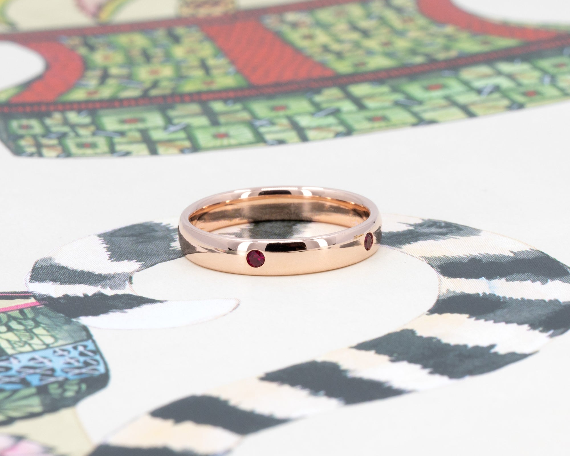 Subtle dome wedding band in rose gold with vivid pigeon blood rubies, photographed on a pastel backdrop by Bena Jewelry.