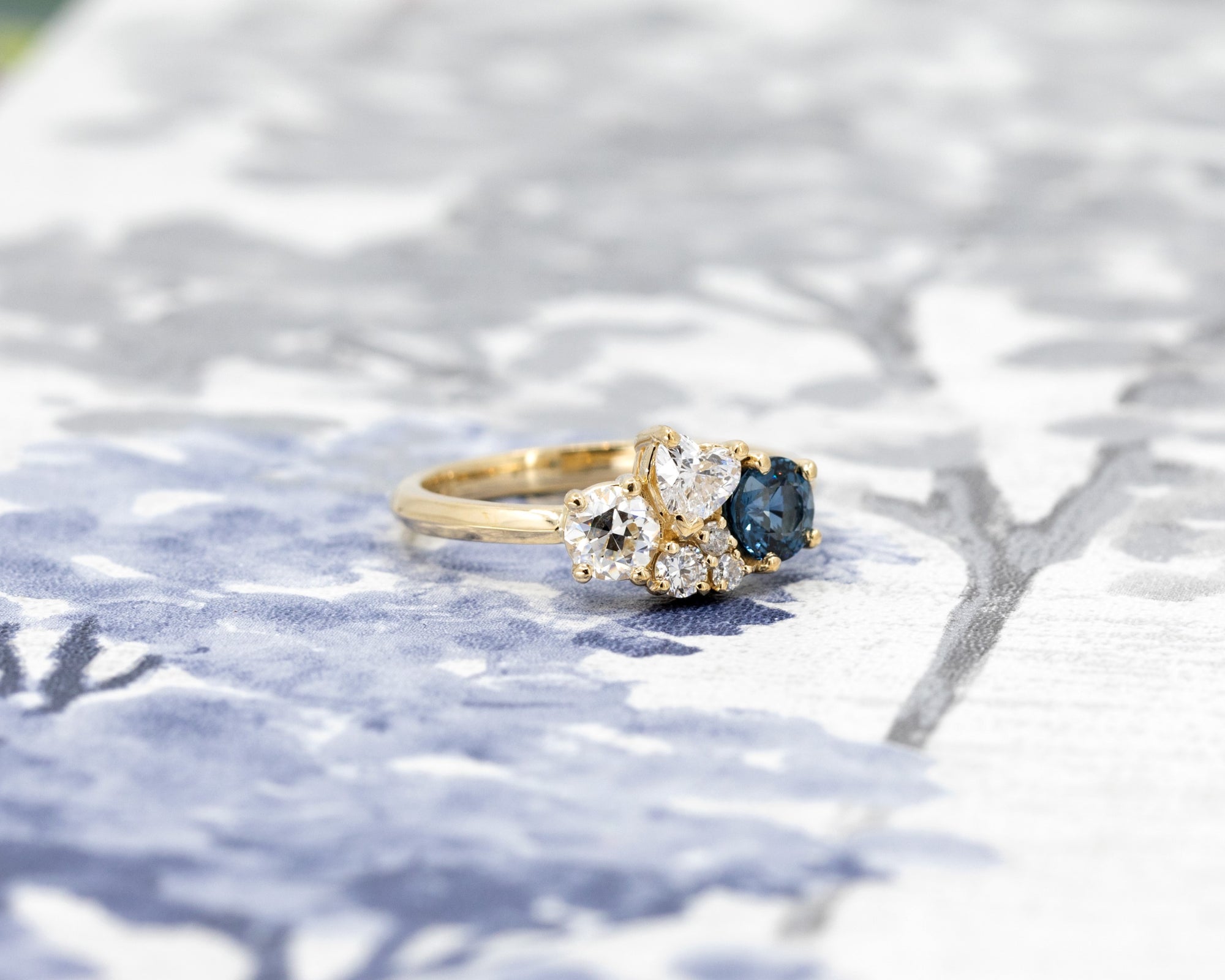 Custom blue spinel and diamond ring in yellow gold, showcased against a light blue background, designed by Bena Jewelry.