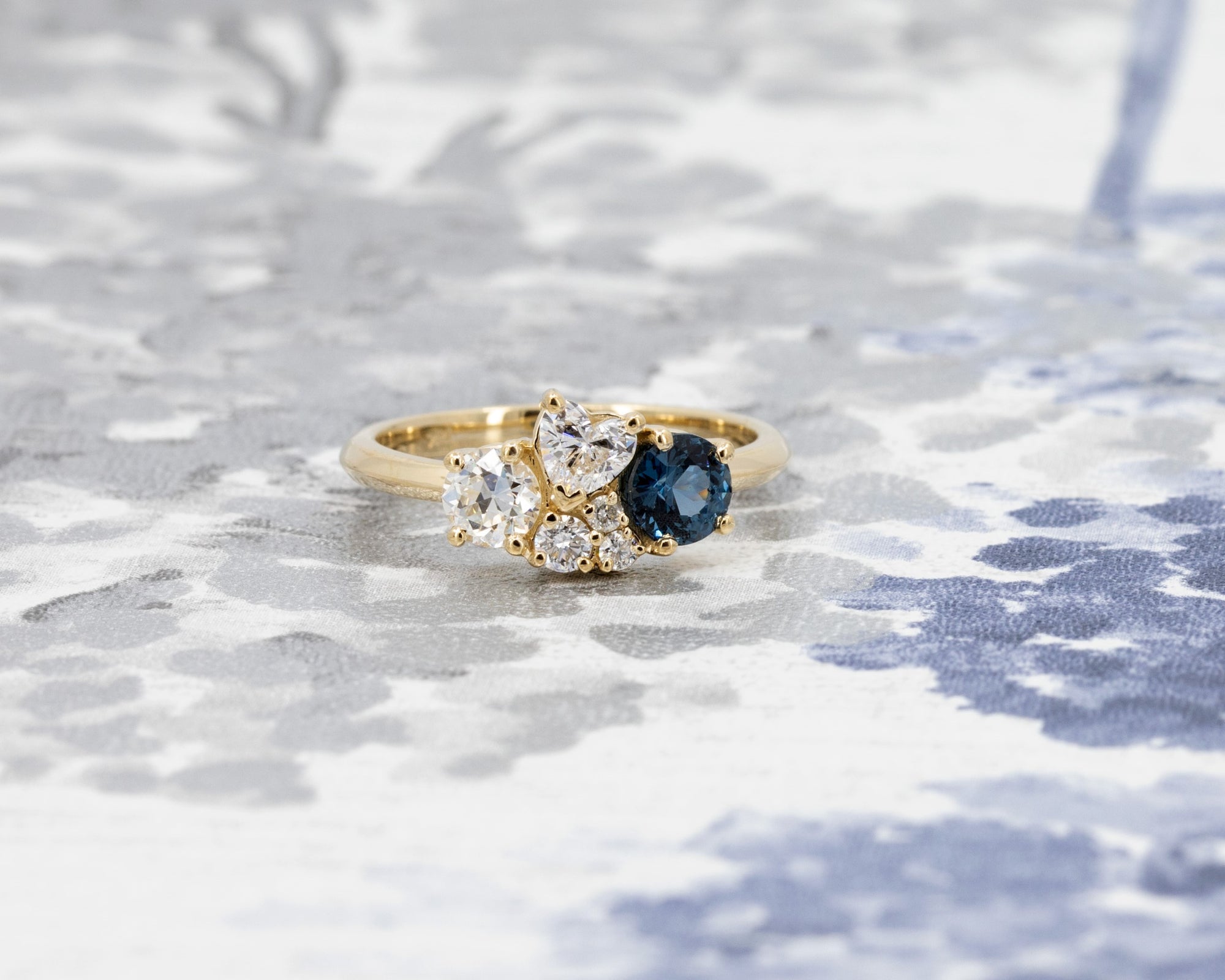 Bespoke fine jewelry featuring a blue spinel and diamonds in yellow gold, styled on a pale blue background.