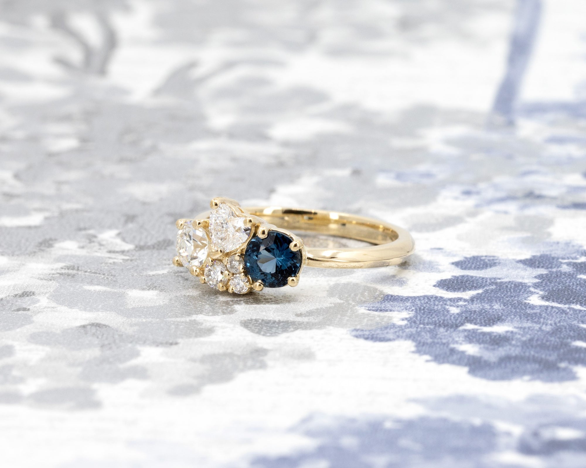 Artistic presentation of a blue spinel and diamond ring in yellow gold, designed by Bena Jewelry, set on a light blue backdrop.