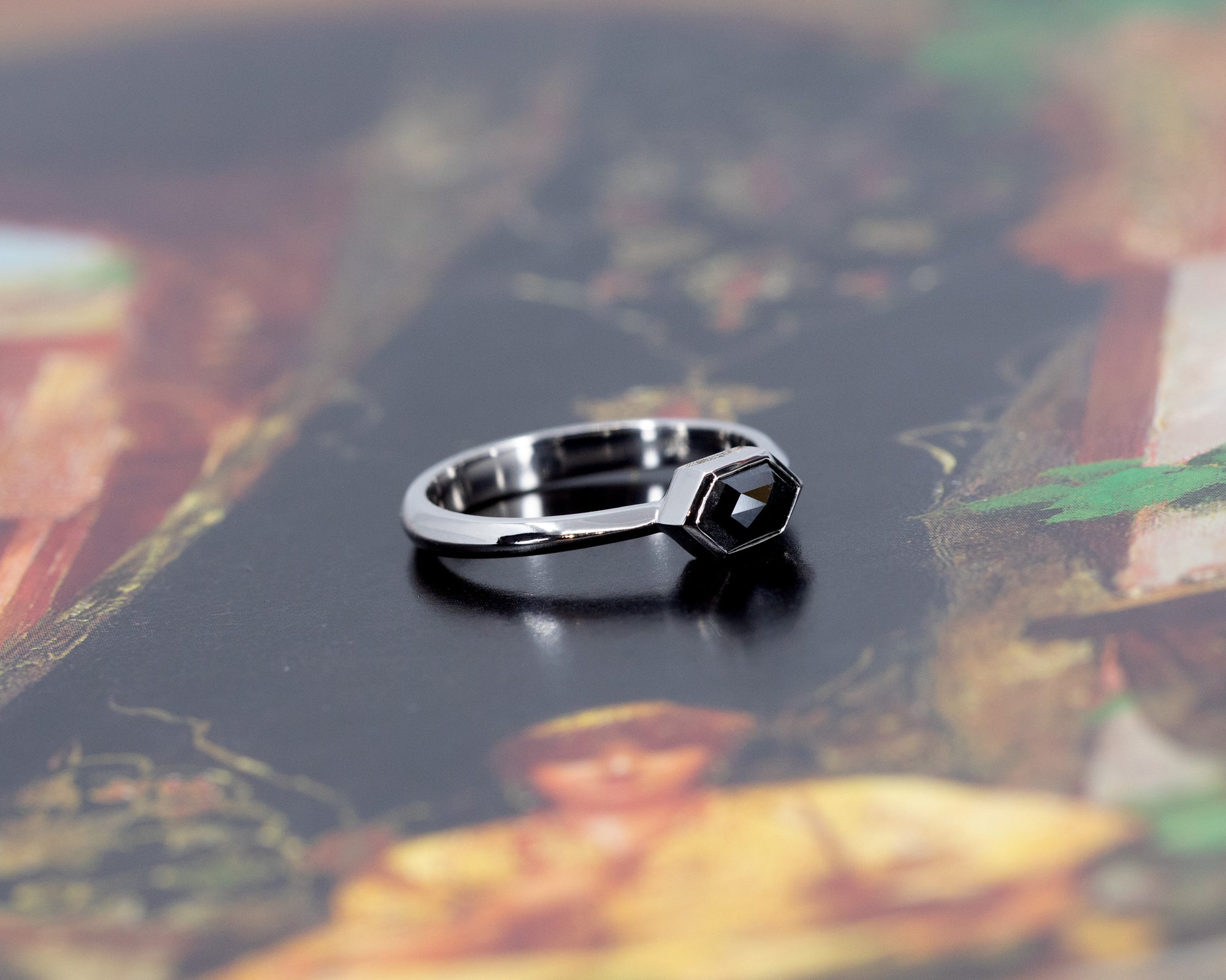 Artistic presentation of a hexagonal black diamond ring in white gold on a soft gradient background, emphasizing the sleek bezel setting and unique design by Bena Jewelry in Montreal, Canada.