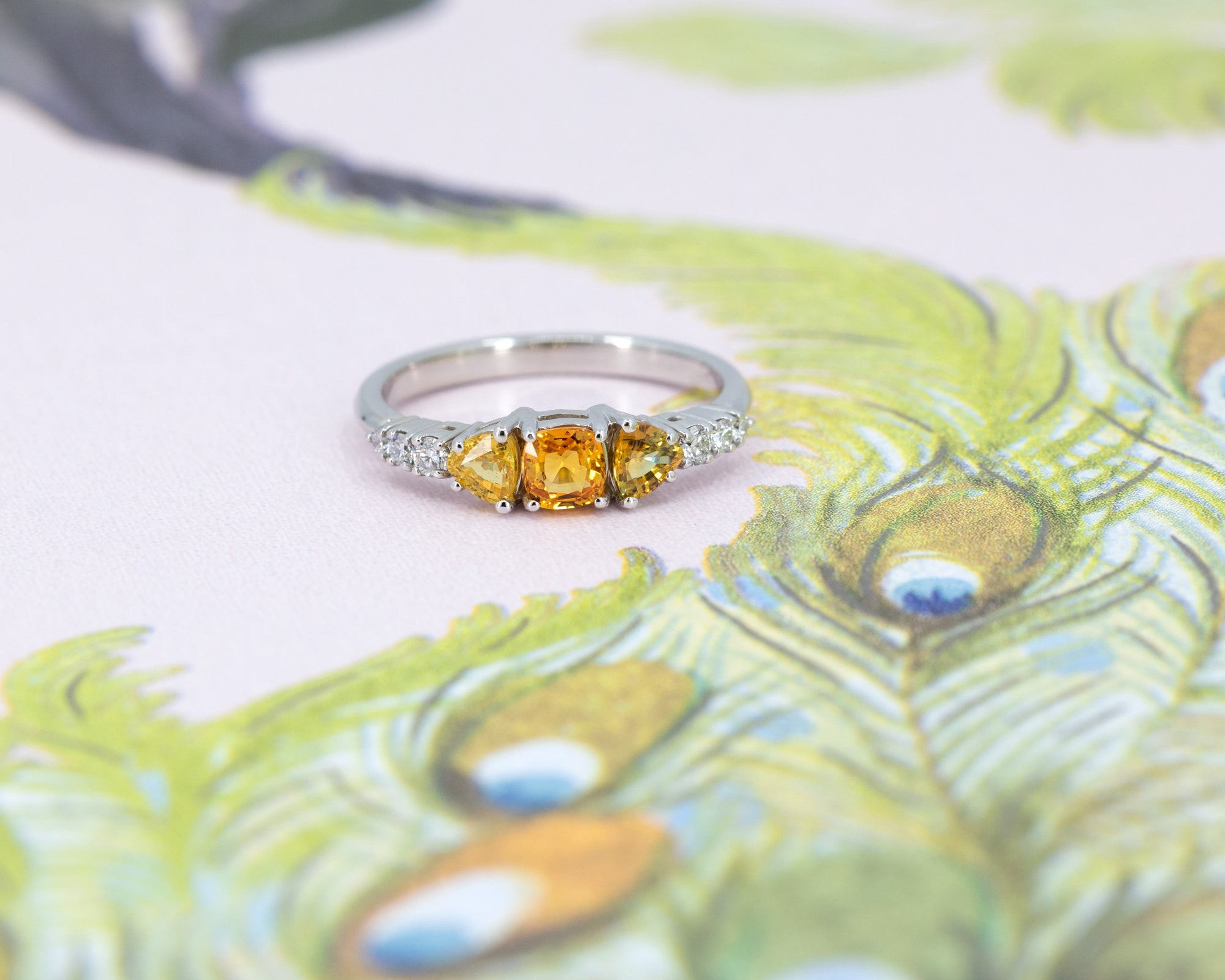 An engagement ring in white gold with three orange sapphires and 5 small white brilliant diamonds si photographed on a stylized illustration of a peacock.