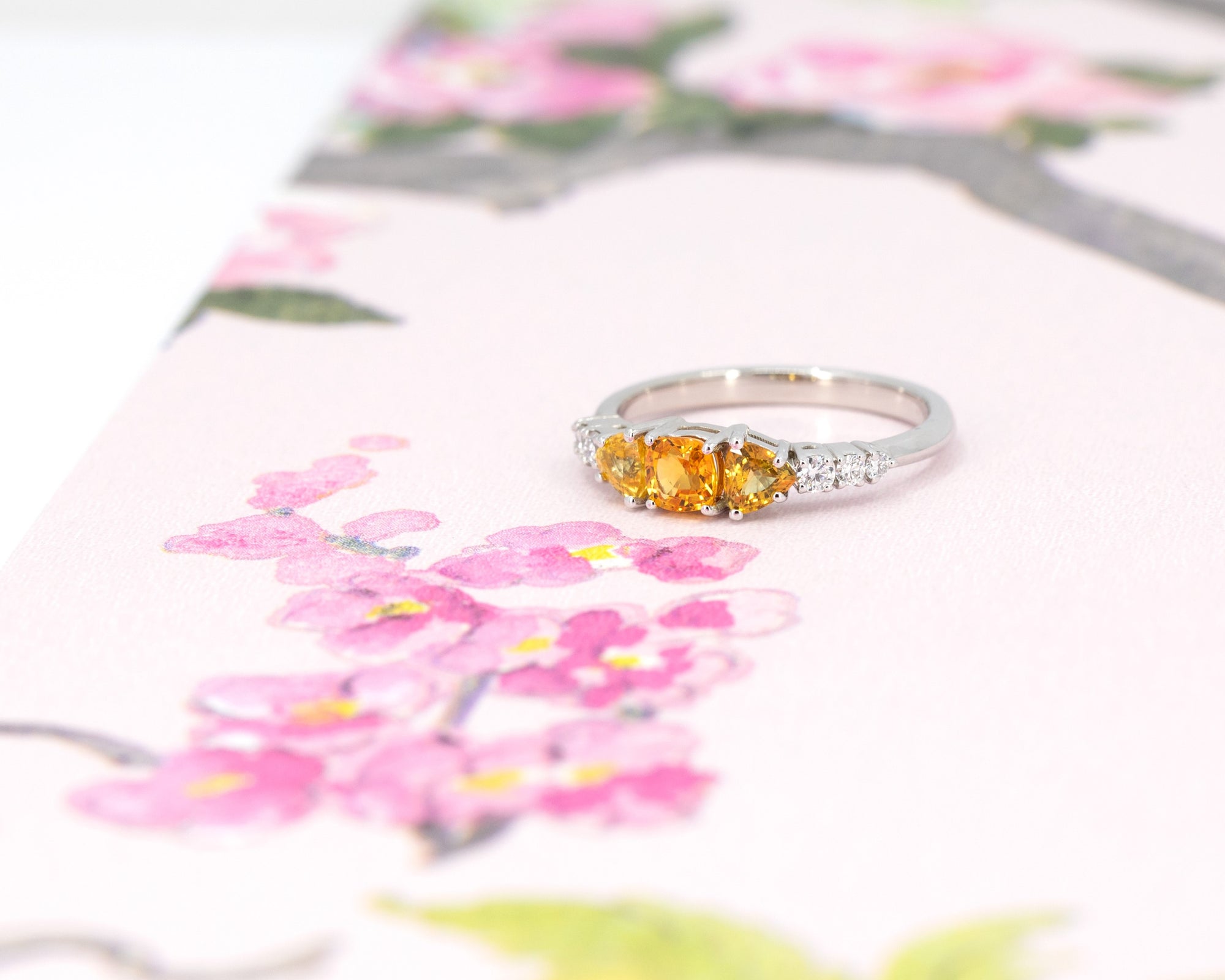 A colorful classic piece of jewelry featuring 3 orange sapphires and 5 extra brilliant white diamonds is photographed on a watercolor image of flowers. This ring was designed by Bena Jewelry, an independent jewellery brand in Montreal, Canada.