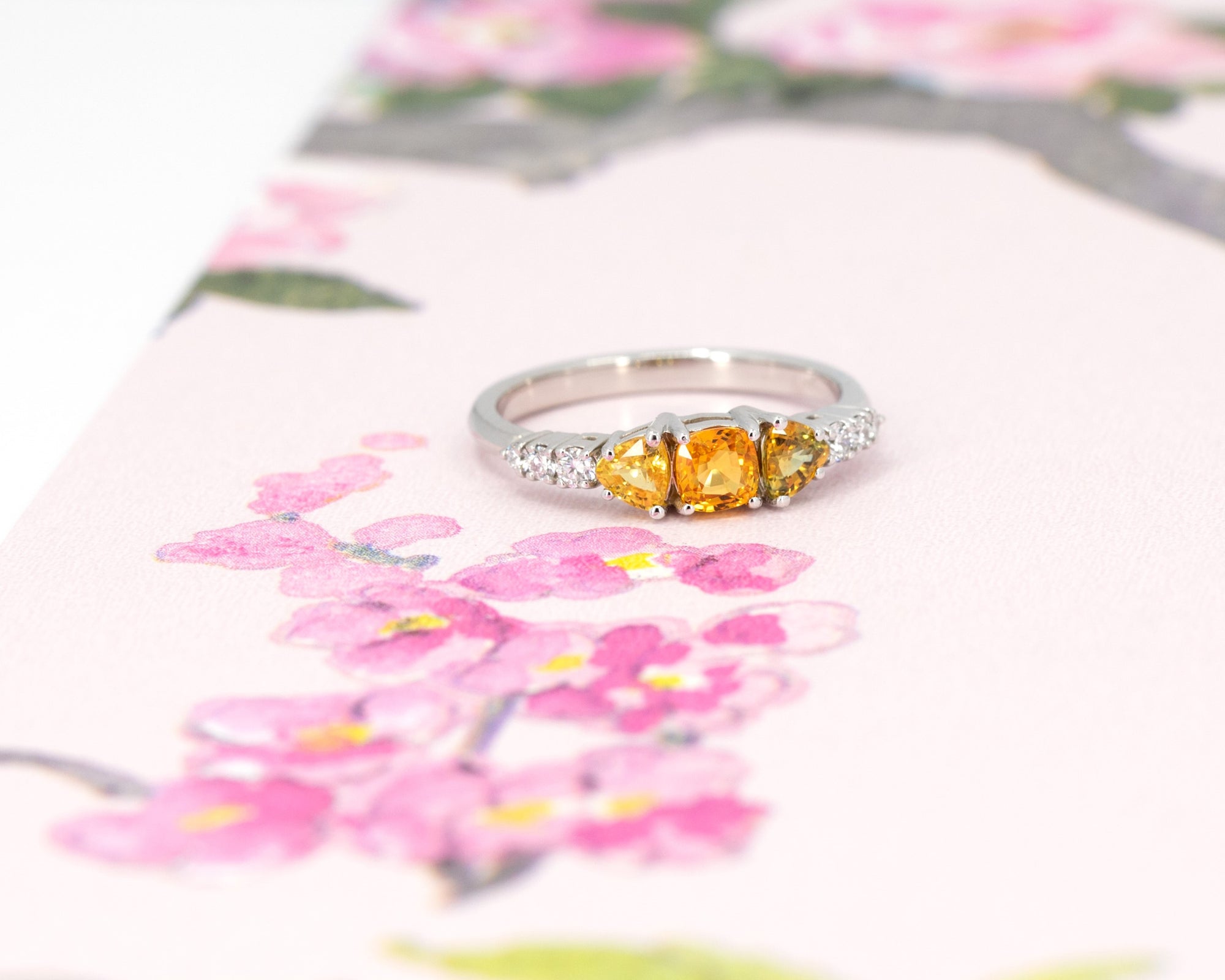 A colorful classic piece of jewelry in white gold featuring 3 orange sapphires and 5 extra brilliant white diamonds is photographed on a watercolor image of flowers. This ring was designed by Bena Jewelry, an independent jewellery brand in Montreal, Canada.
