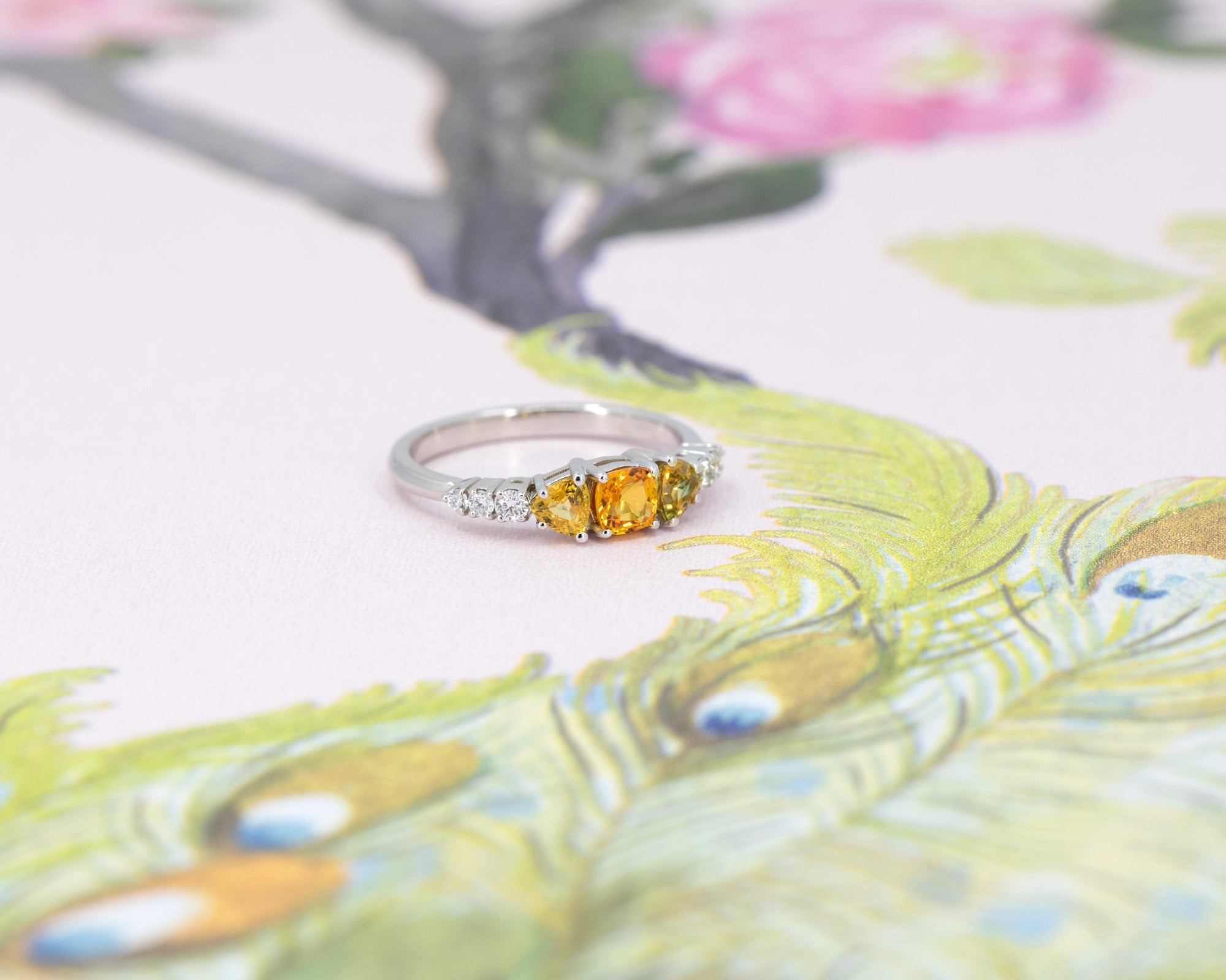 An engagement ring in white gold with three orange sapphires and 5 small white brilliant diamonds si photographed on a stylized illustration of a peacock. This fine piece of jewellery was designed by Bena Jewelry in Montreal, Canada.
