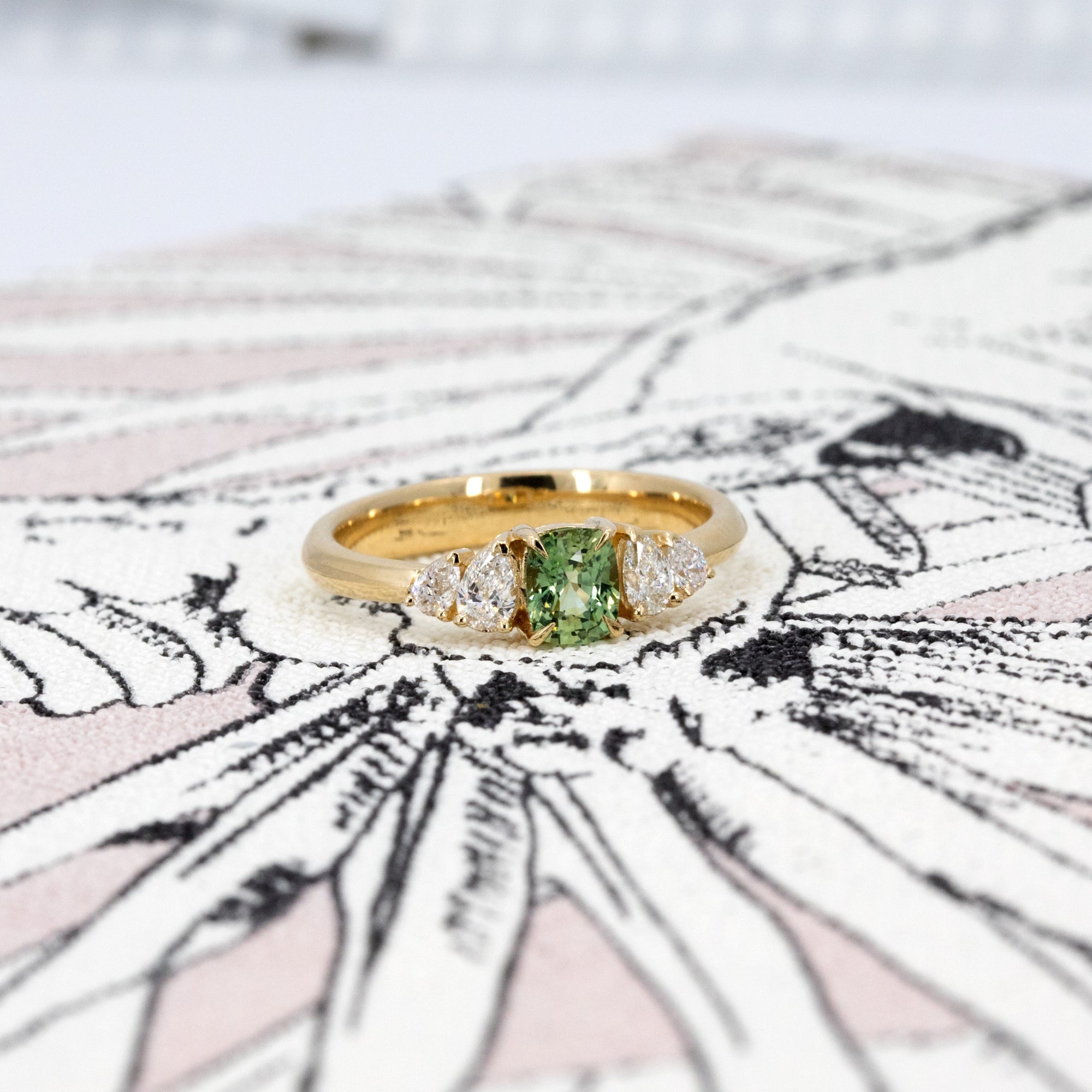 Green sapphire cushion gemstone ring with lab-created pear diamond accents in a yellow gold setting. Designed by Bena Jewelry in Montreal, Canada.