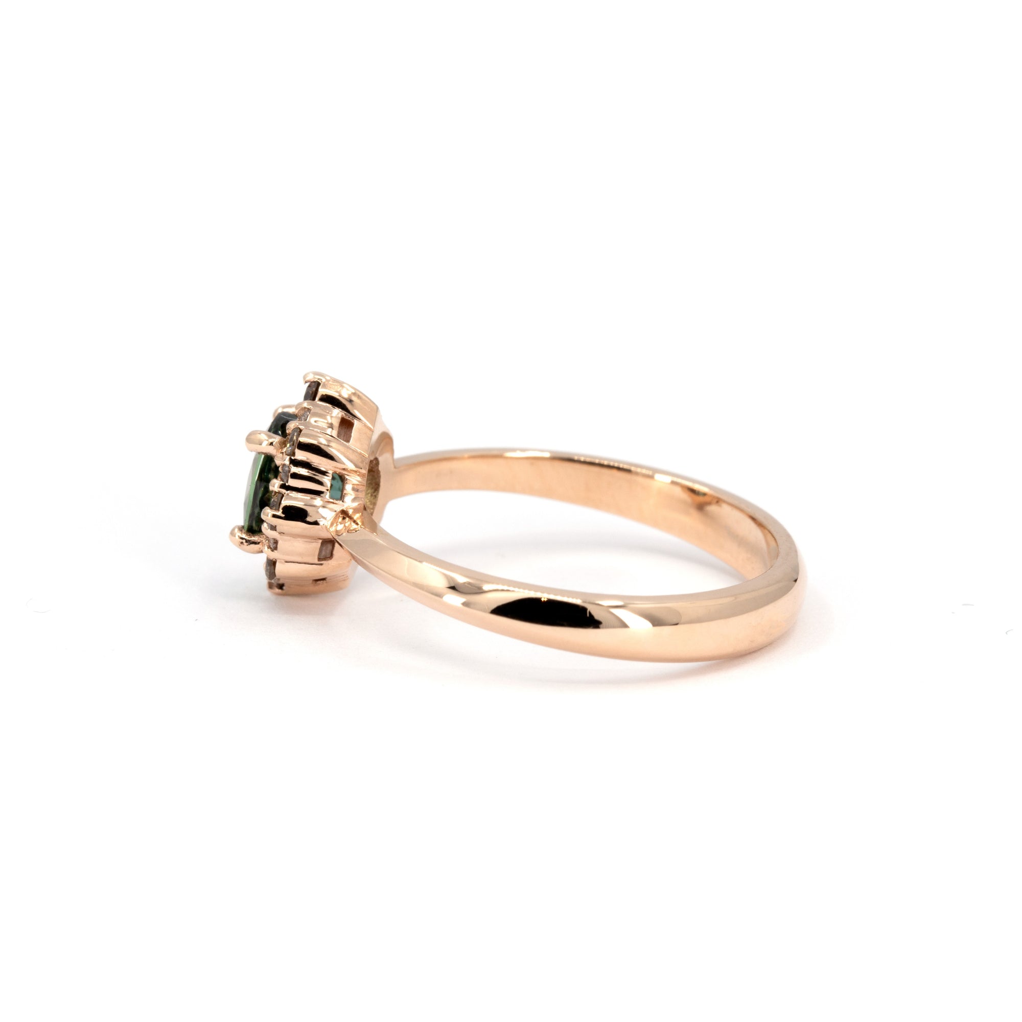 Side view of the Aura ring by Bena Jewelry with a deep teal sapphire and diamond halo, set in rose gold, photographed on a white surface.