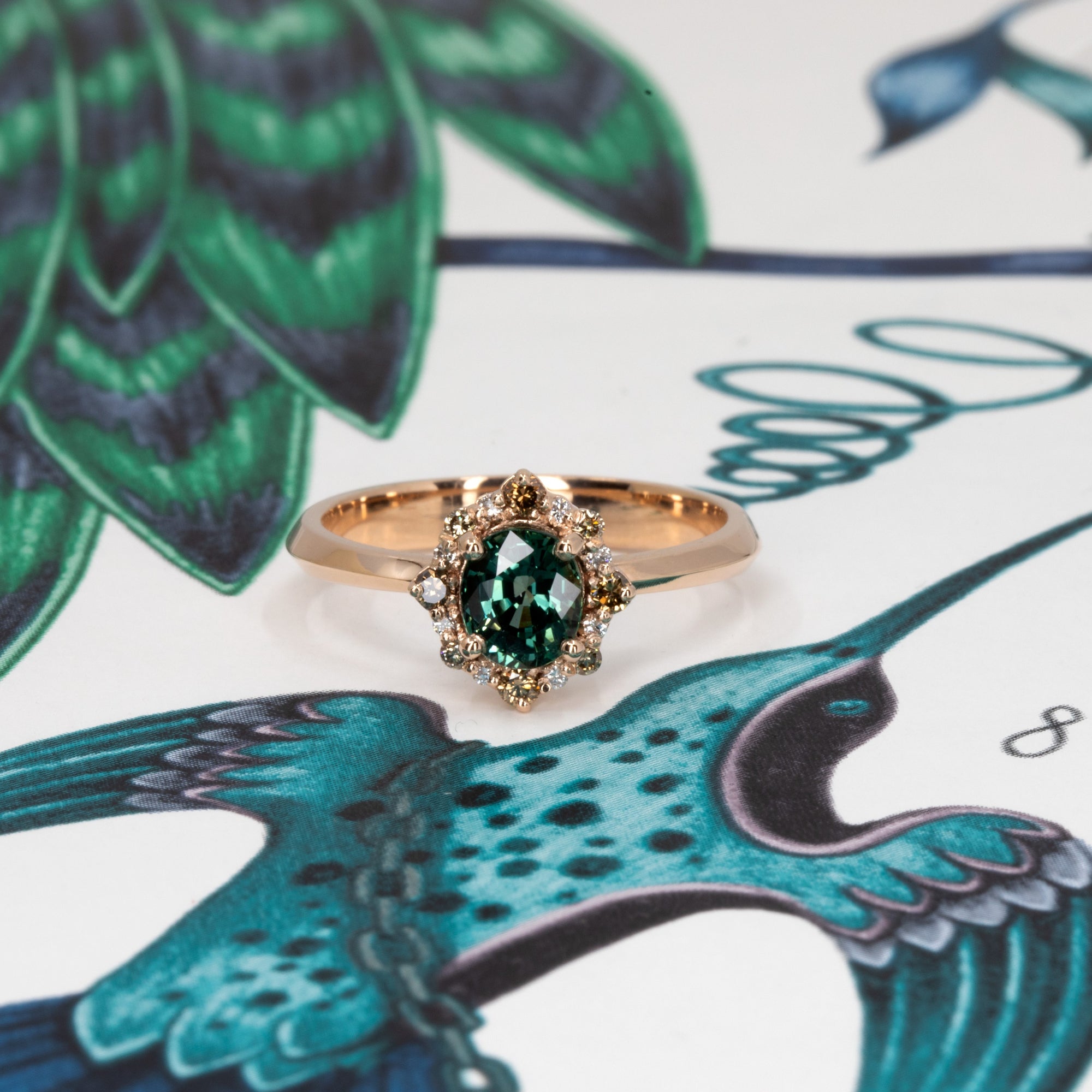 Elegant rose gold ring with teal sapphire center and sparkling diamond halo, designed by Bena Jewelry, captured on a warm-toned background.