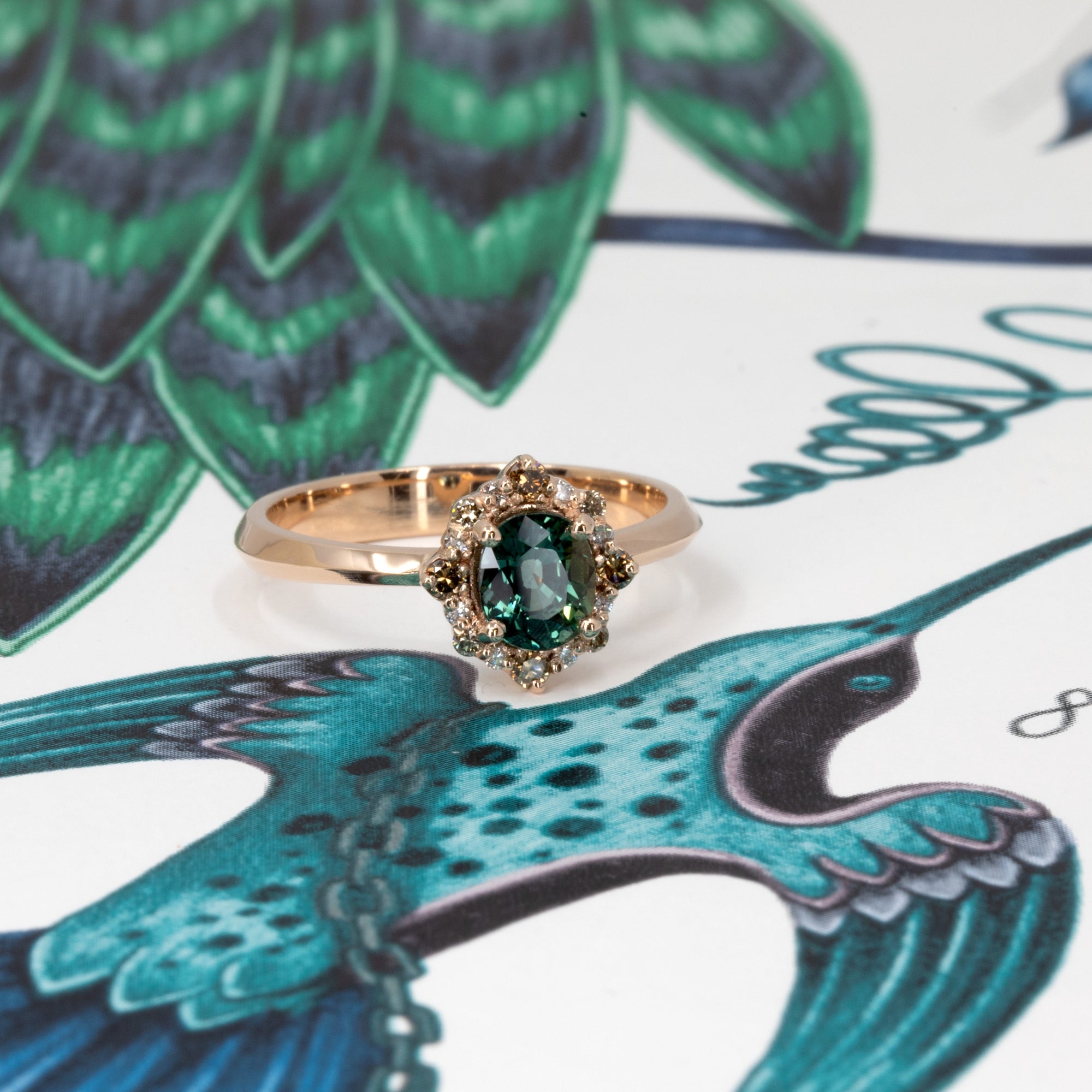 Teal sapphire and diamond halo ring in rose gold by Bena Jewelry, photographed on a soft pink-beige background in Montreal.