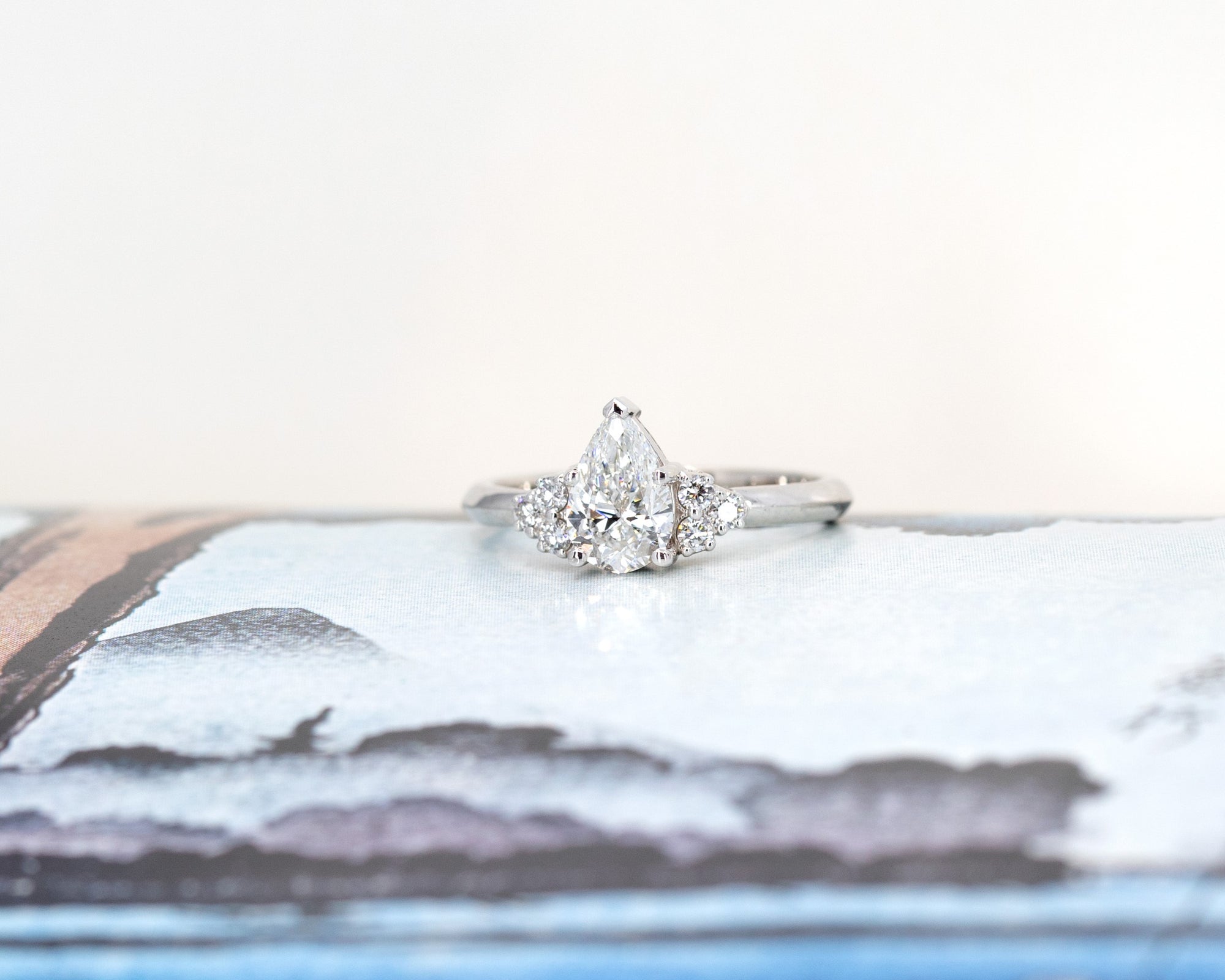 Pear-shaped diamond engagement ring in white gold with side diamonds, showcased on a soft pastel background. Crafted by Bena Jewelry in Montreal.