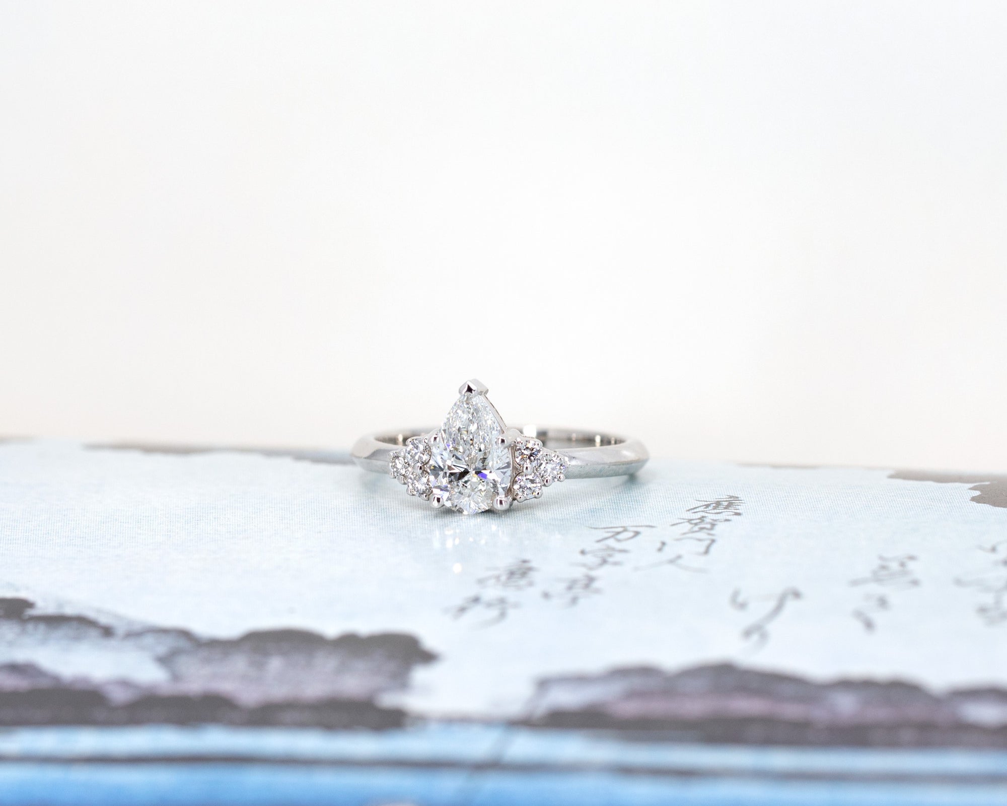 Custom white gold ring with a pear-shaped diamond and side accents, photographed on a colorful background. Designed by Bena Jewelry in Montreal, Canada.