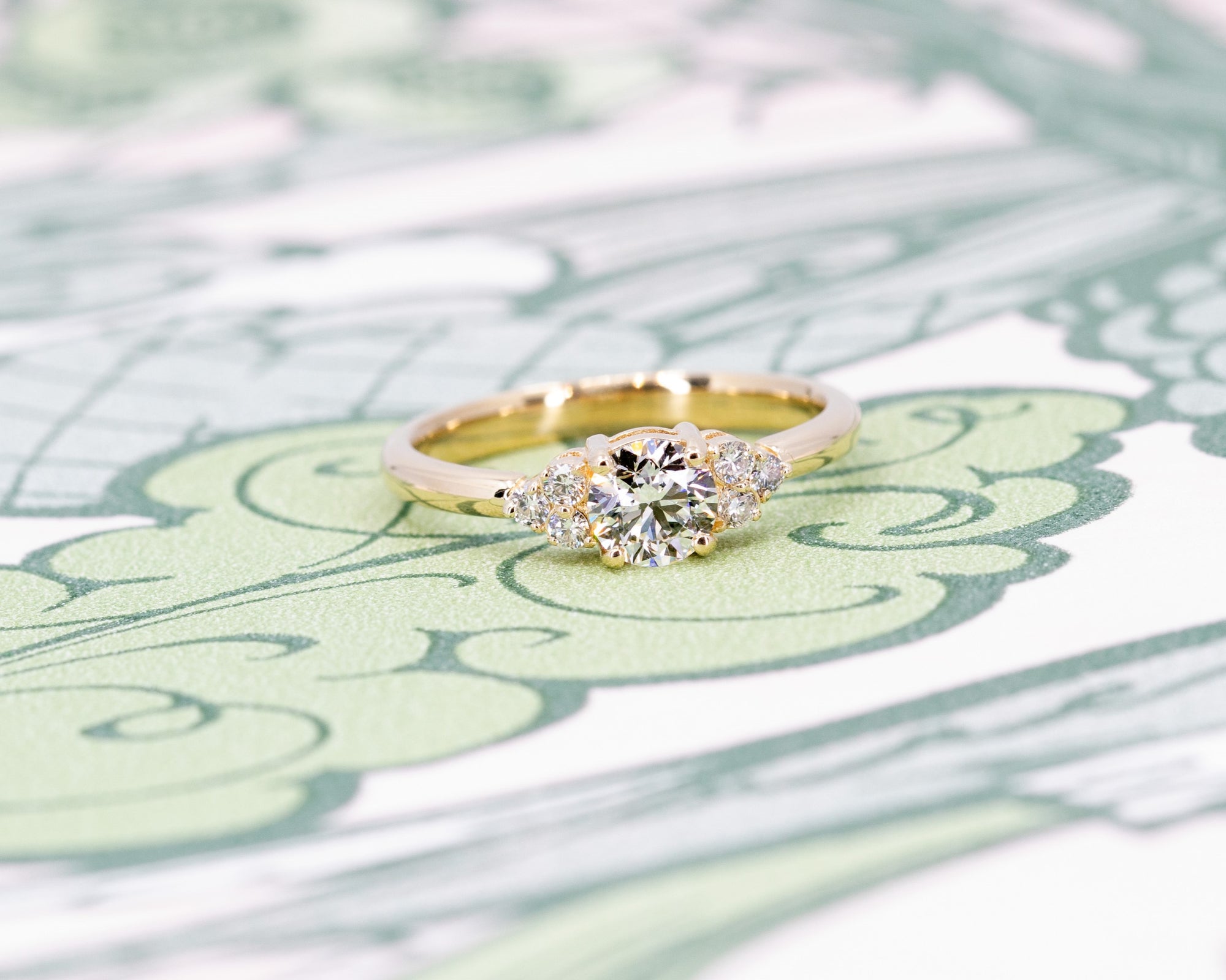 Yellow gold diamond ring with a timeless design, featuring a round center stone and side accents, photographed on a pastel backdrop by Bena Jewelry.