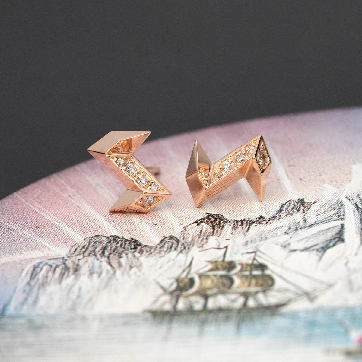 A pair of earrings in the shape of a lightning bolt, but with a unique style as they are made of rose gold with diamonds, and the earrings are made with several facets in volume that reflect light in several directions. This fine jewelry was designed and created in Montreal by local brand Bena Jewelry. The stud earrings are photographed on an artistic image featuring a sailboat and mountains. 