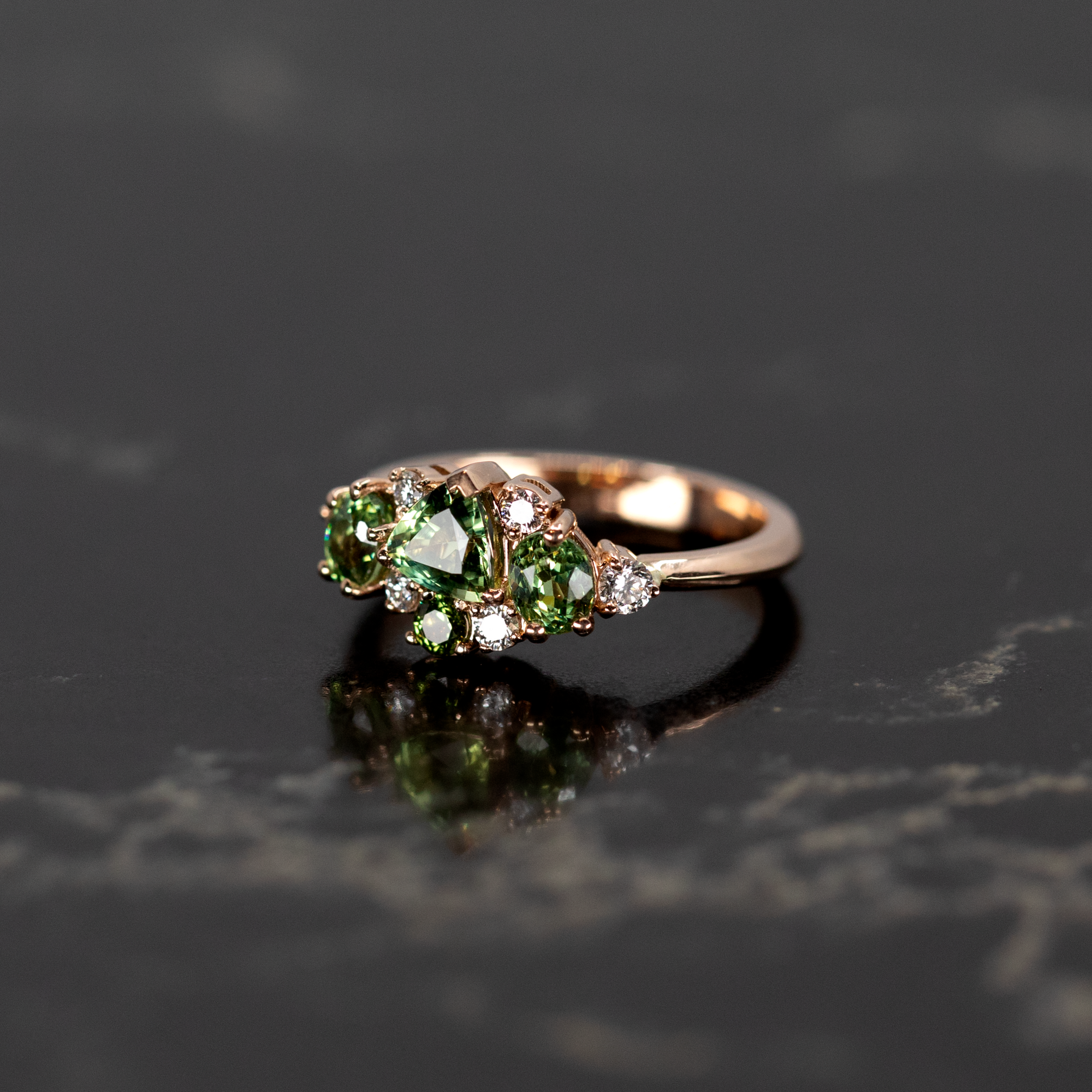 Vibrant green sapphire and demantoid garnet ring in rose gold, displayed on a dark backdrop. Designed by Bena Jewelry in Montreal.