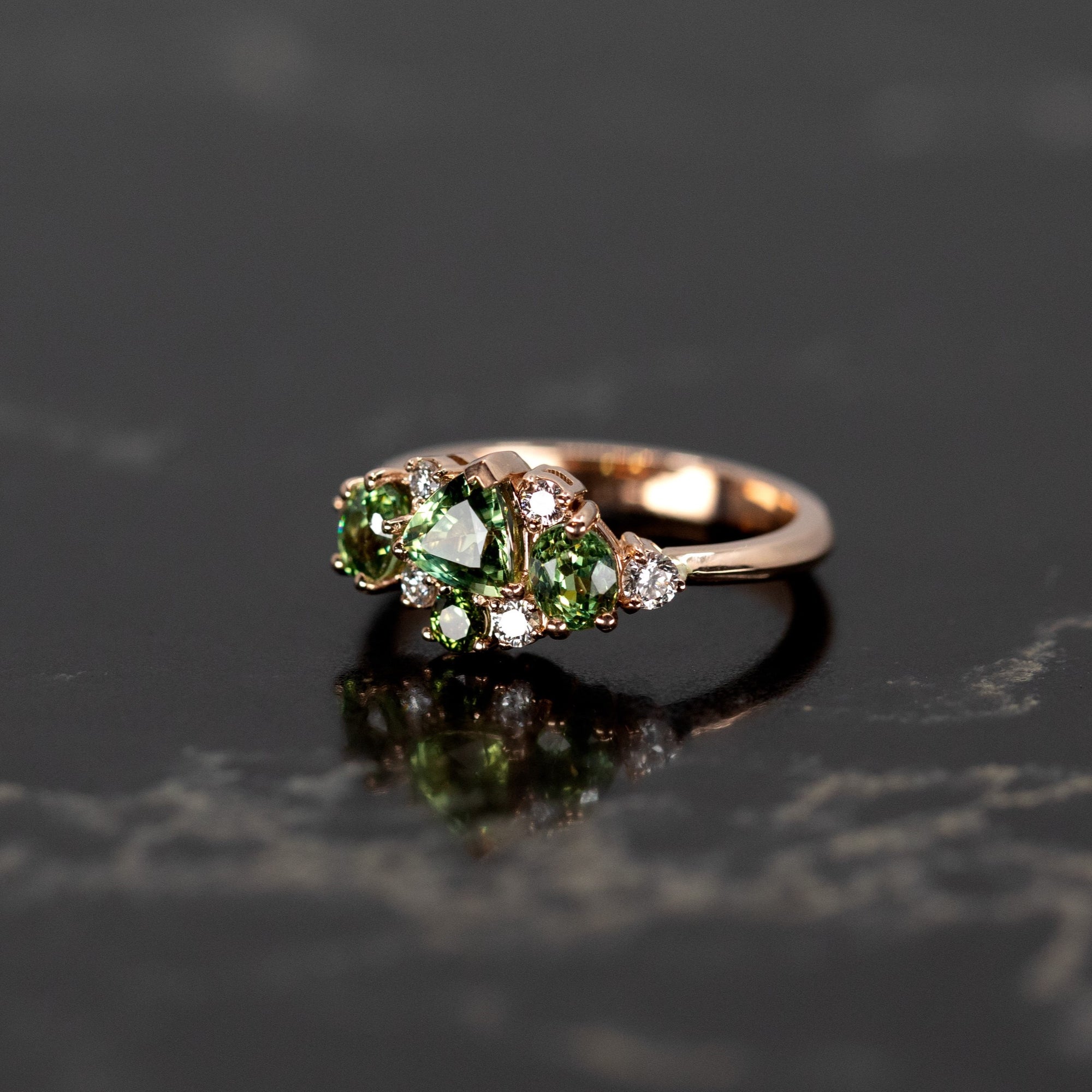 Unique rose gold ring with green gemstones, including sapphire and garnet, shown against a dark background. Made by Bena Jewelry, Montreal, Canada.