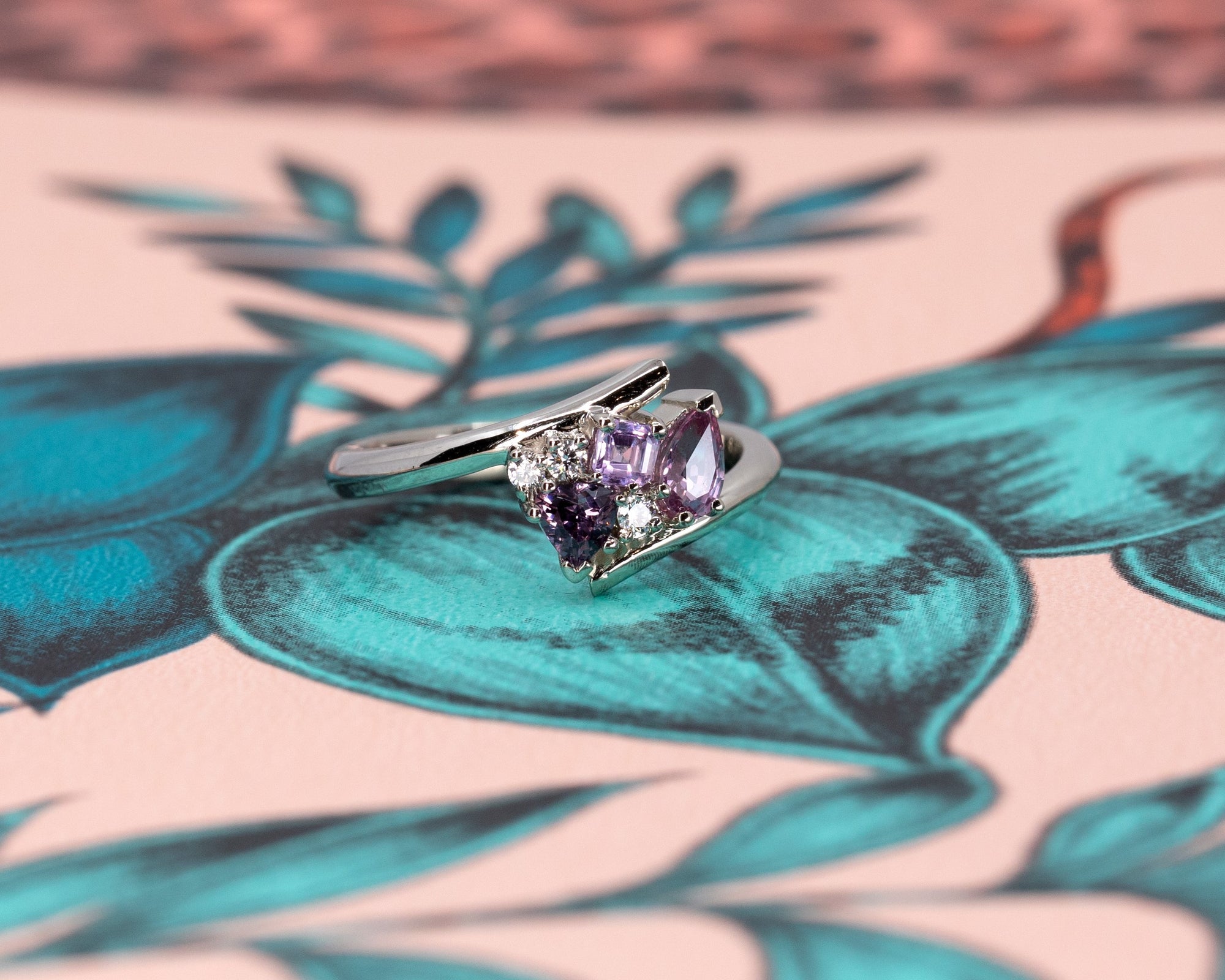 Close-up of Bena Jewelry's Kink Avalanche white gold ring, highlighting the vivid purple and pink sapphires in various cuts and sparkling diamonds, set against a colorful backdrop.