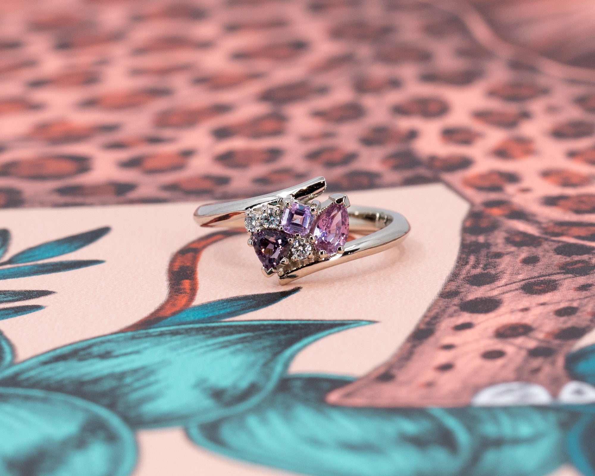 Side view of the Kink Avalanche white gold ring's dynamic twist band design, featuring purple and pink sapphires and diamonds, on a colorful background.