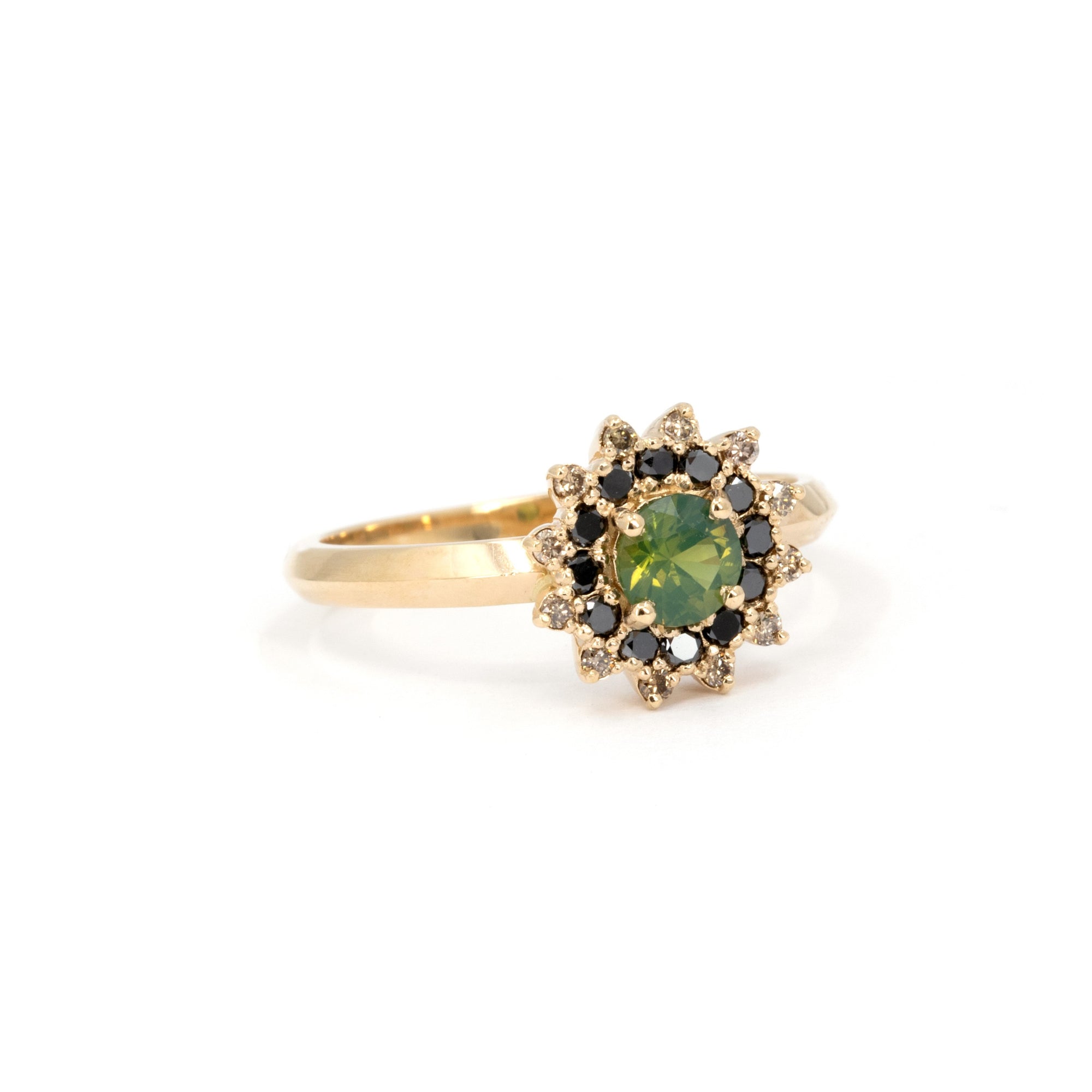 Close-up of a handmade green sapphire ring by Bena Jewelry, with black and brown diamond halo and faceted yellow gold band, photographed on white surface.