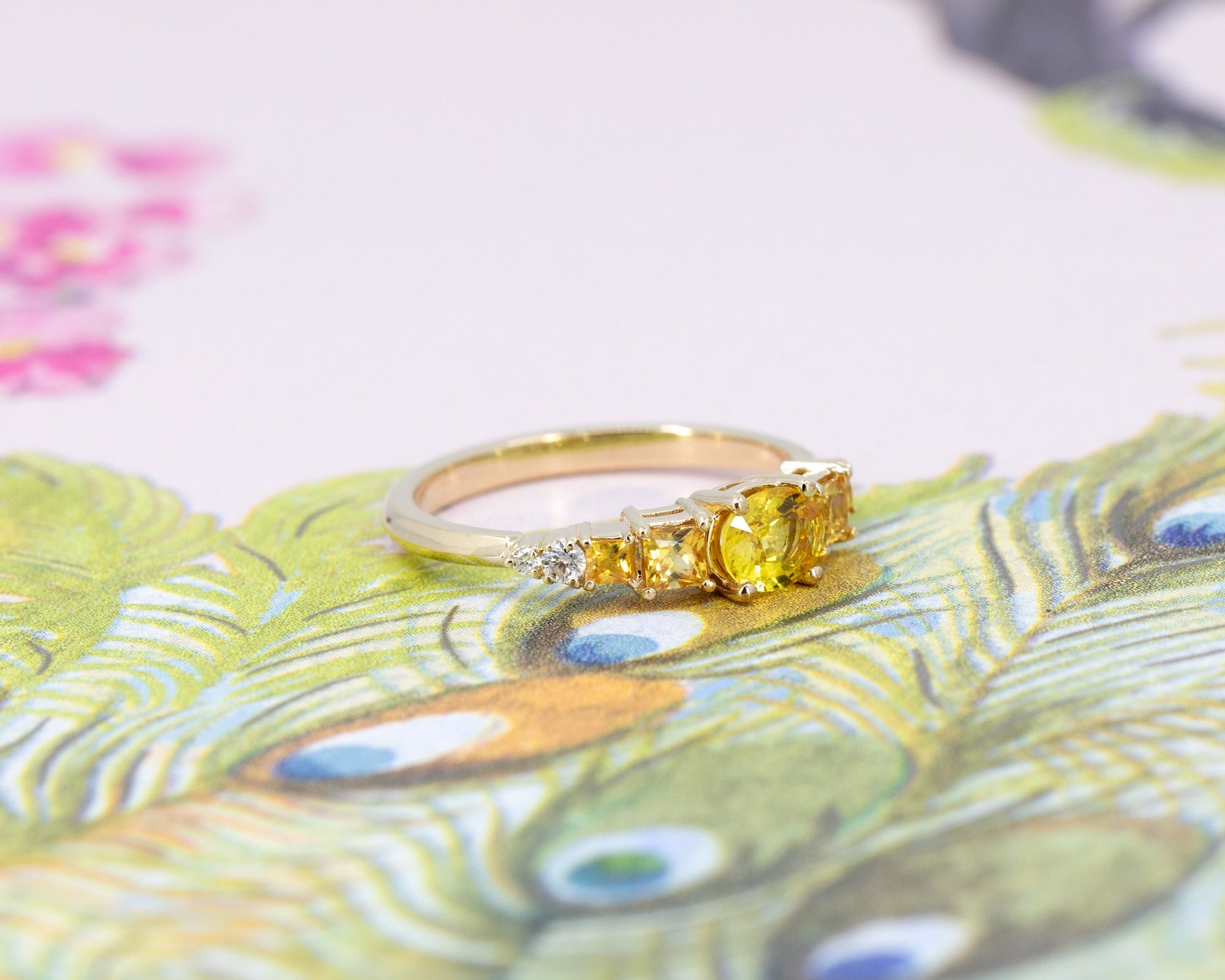 Yellow gold engagement ring featuring a vivid yellow oval sapphire center stone, flanked by orange princess cut sapphires and diamonds, displayed against a floral background.