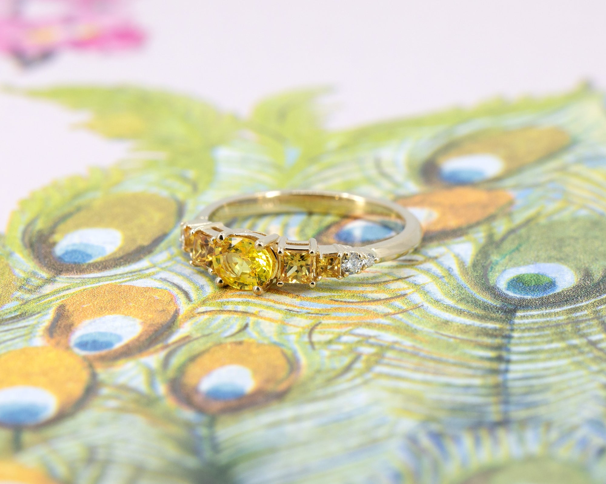 Close-up of the yellow gold engagement ring highlighting the vivid yellow oval sapphire center stone, orange princess cut sapphires, and diamond accents, set against a floral backdrop.