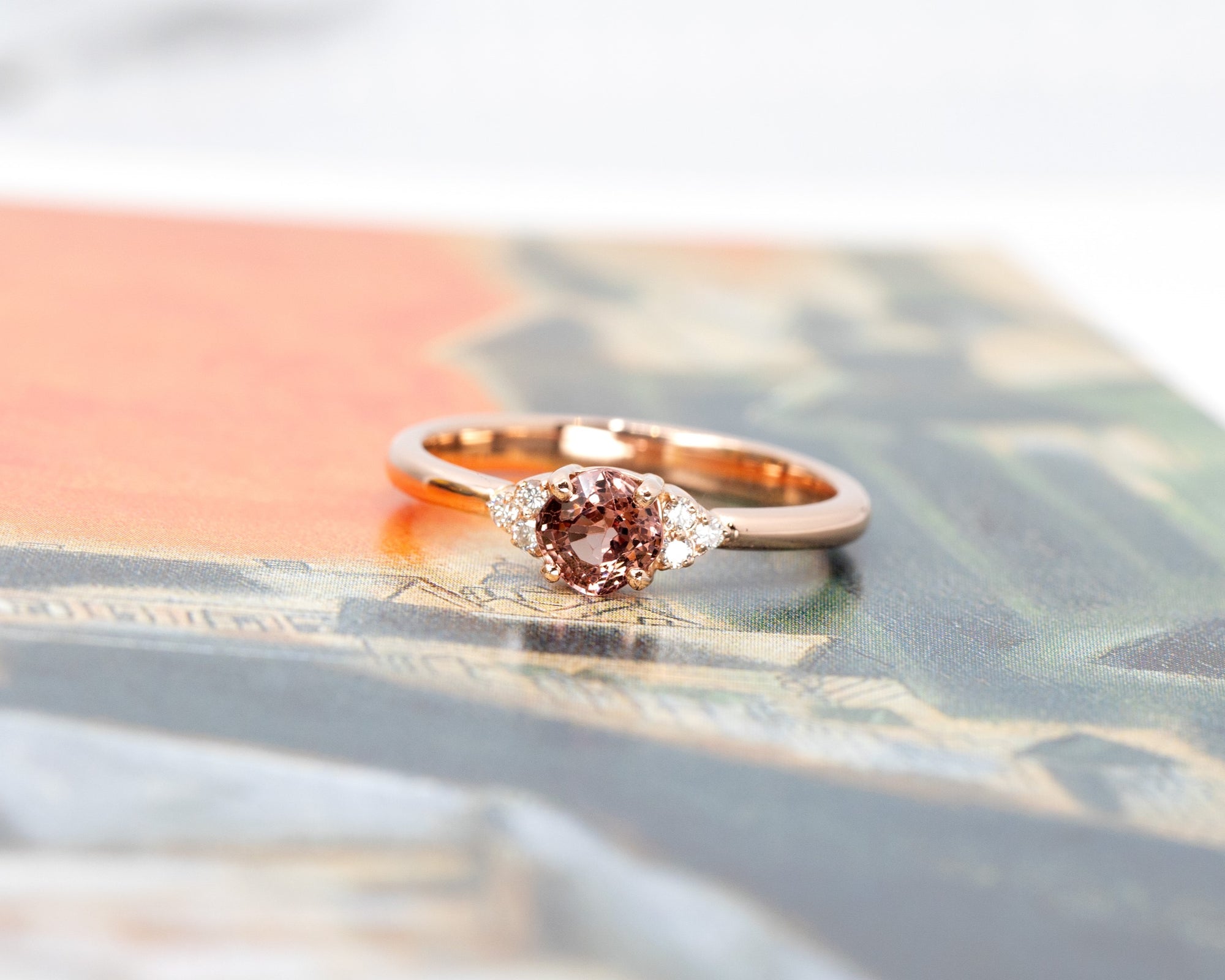 The Désir Peach Spinel Ring set against a warm-toned background, highlighting its cushion-cut gemstone and rose gold setting. A refined creation by Bena Jewelry.