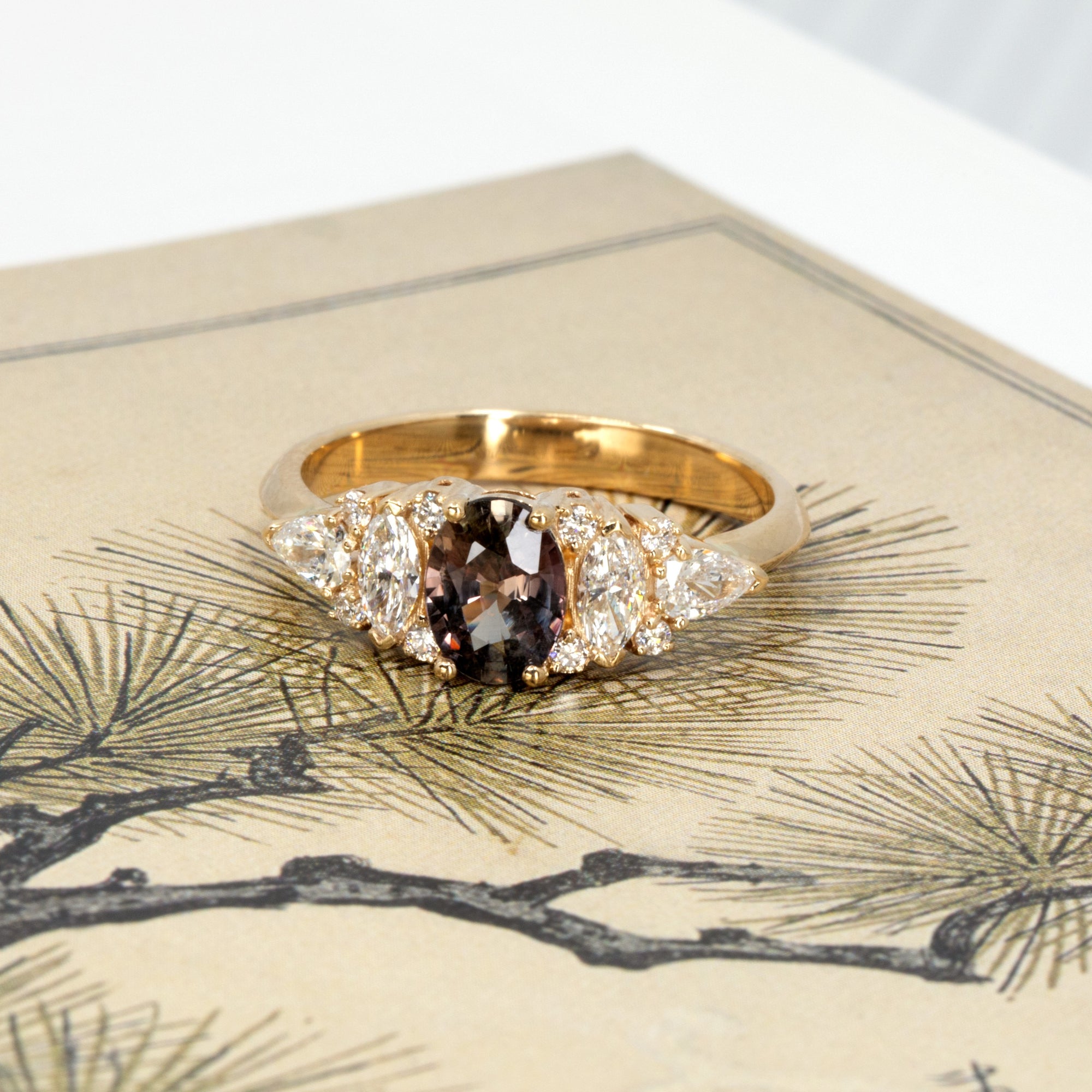Artistic shot of a handcrafted engagement ring with oval peacock sapphire and mixed-cut diamonds, designed by Bena Jewelry in Montreal, set against a moody colored background.
