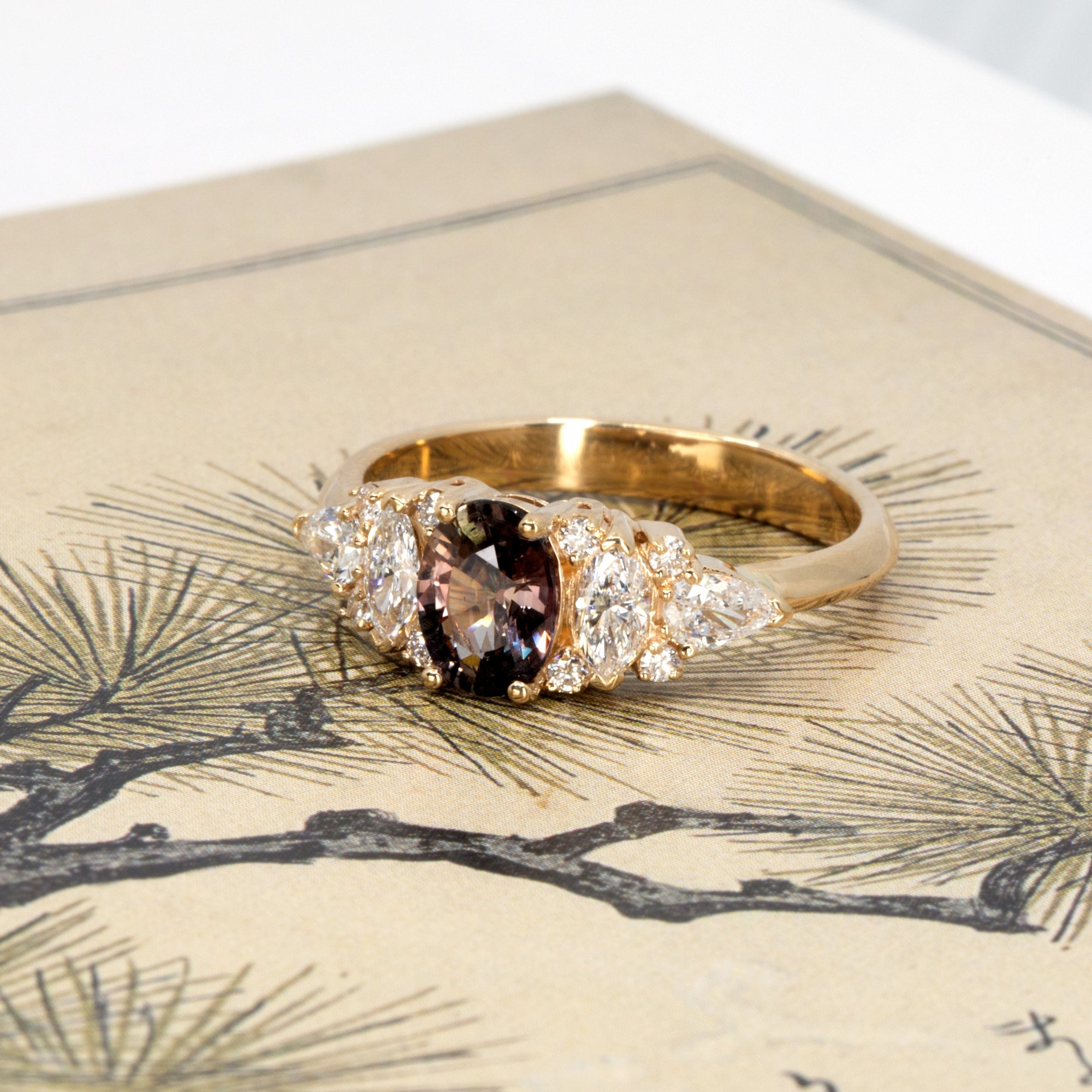 Bold peacock sapphire and diamond ring by Bena Jewelry displayed on a deep-toned background, highlighting the rich hues of the center stone and 14k gold setting.