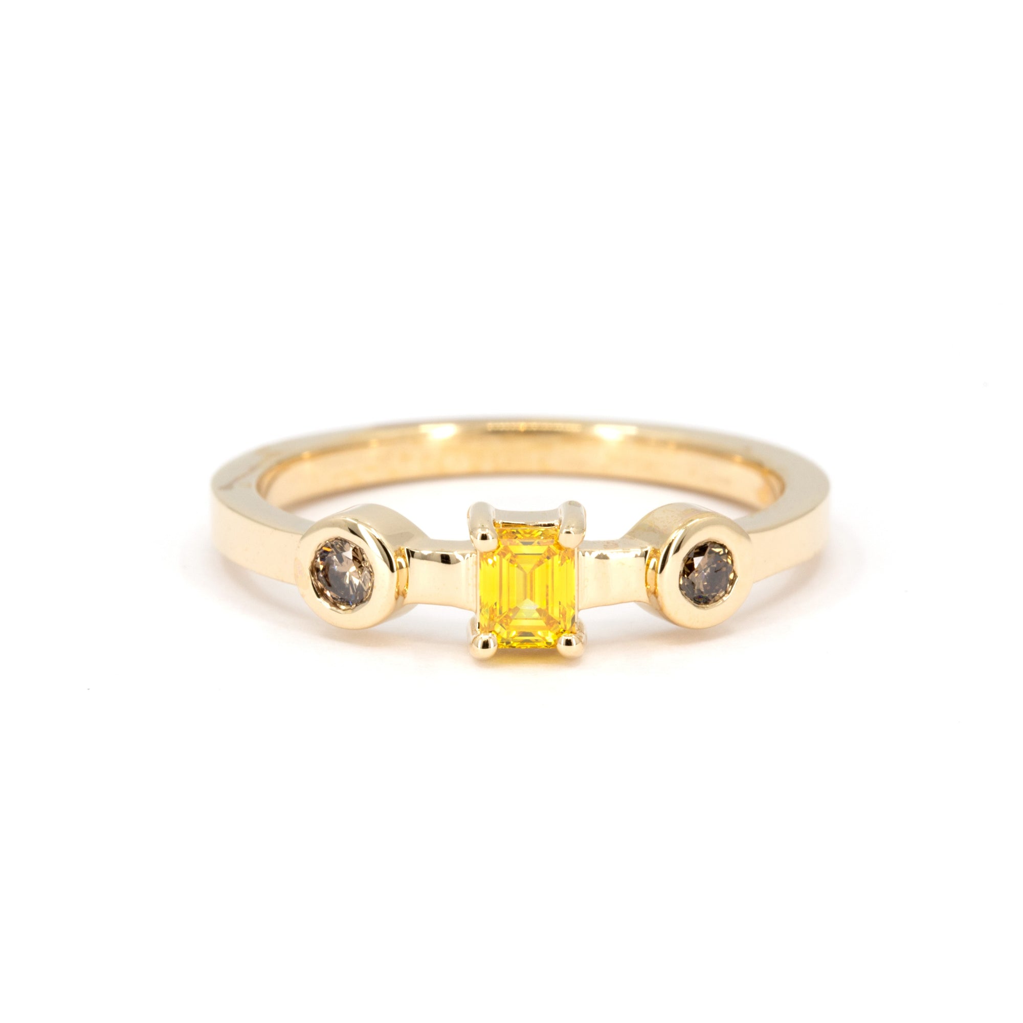 Close-up view of a yellow gold ring featuring an emerald-cut yellow diamond, flanked by two bezel-set natural brown diamonds, against a white background.