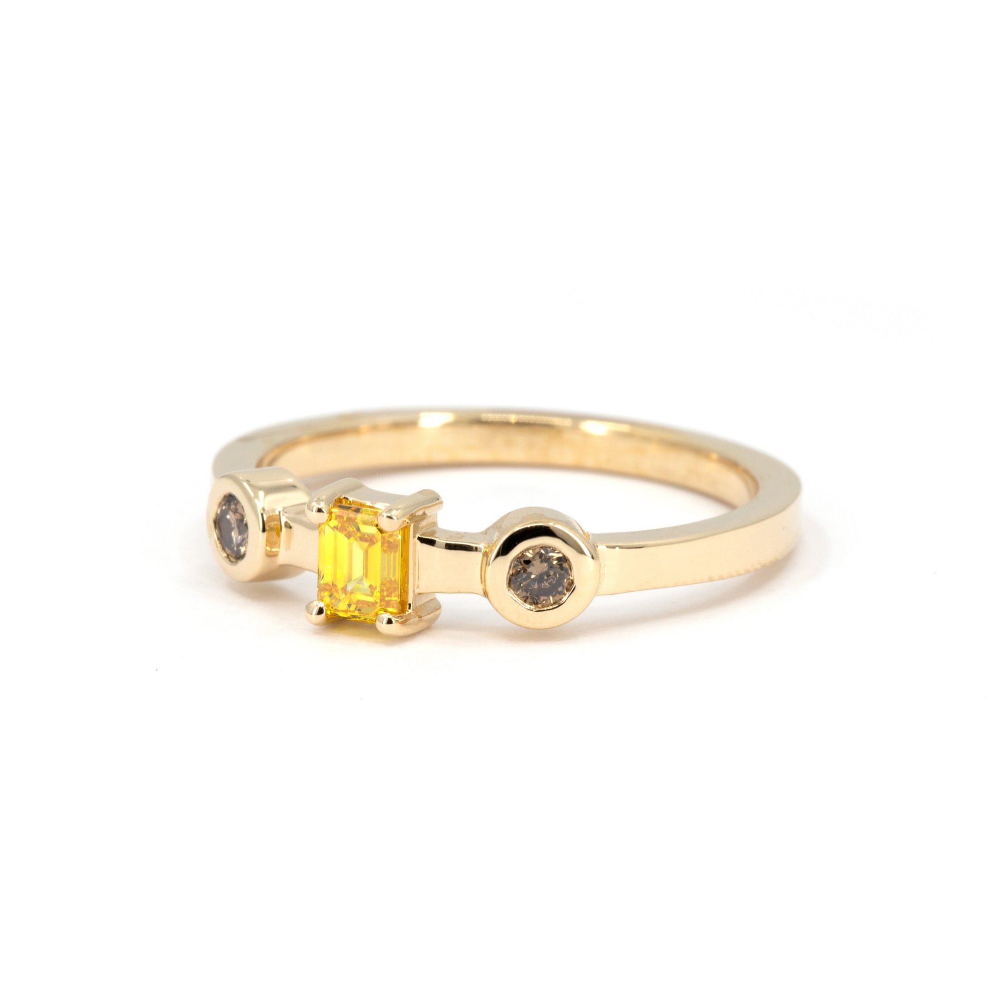 Minimalist shot of a handcrafted yellow gold ring with a central yellow emerald-cut diamond and two side brown diamonds, set in bezel settings, displayed on a white background.
