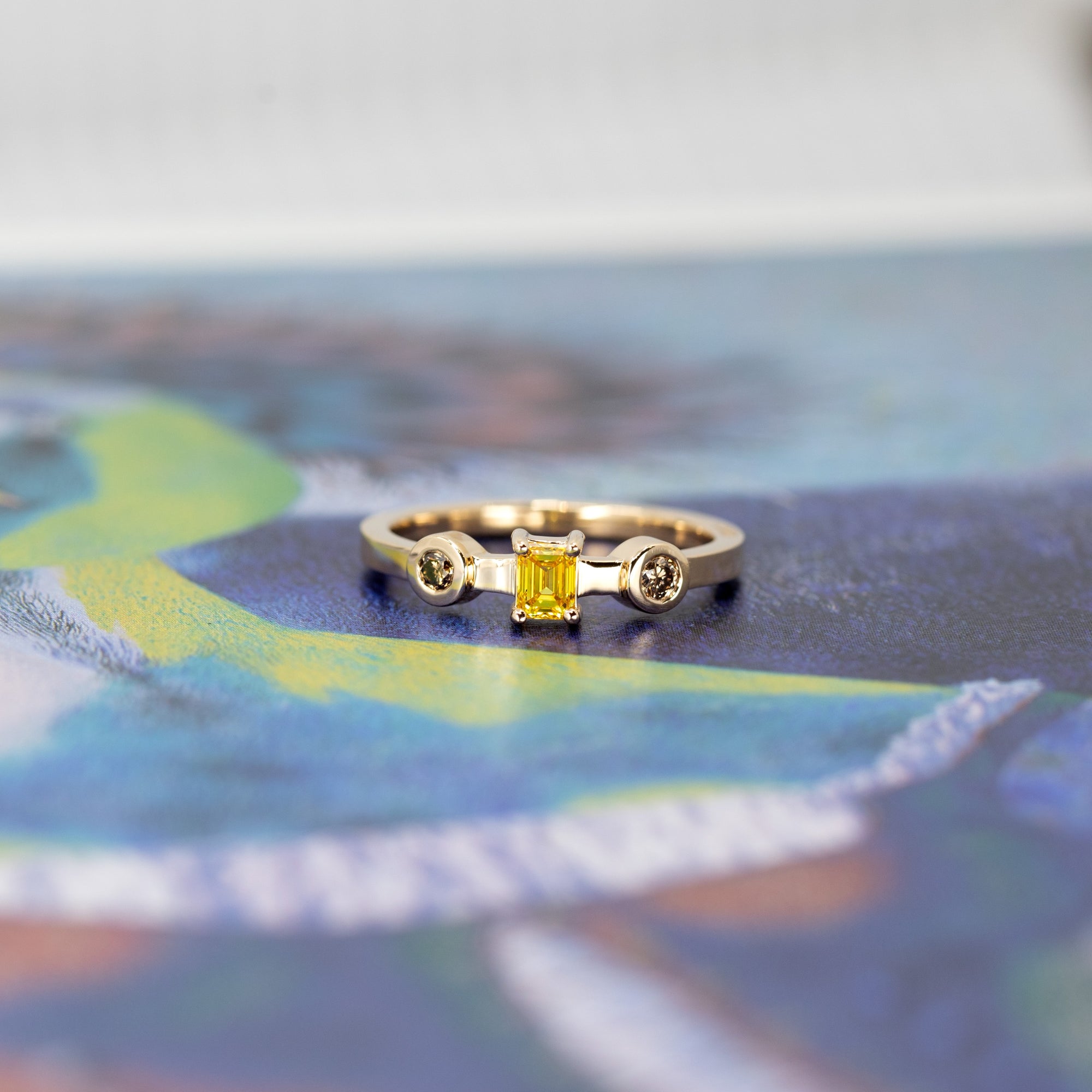 Elegant close-up of a handcrafted gold ring with a yellow diamond centerpiece and brown diamonds, photographed on a contrasting color background.