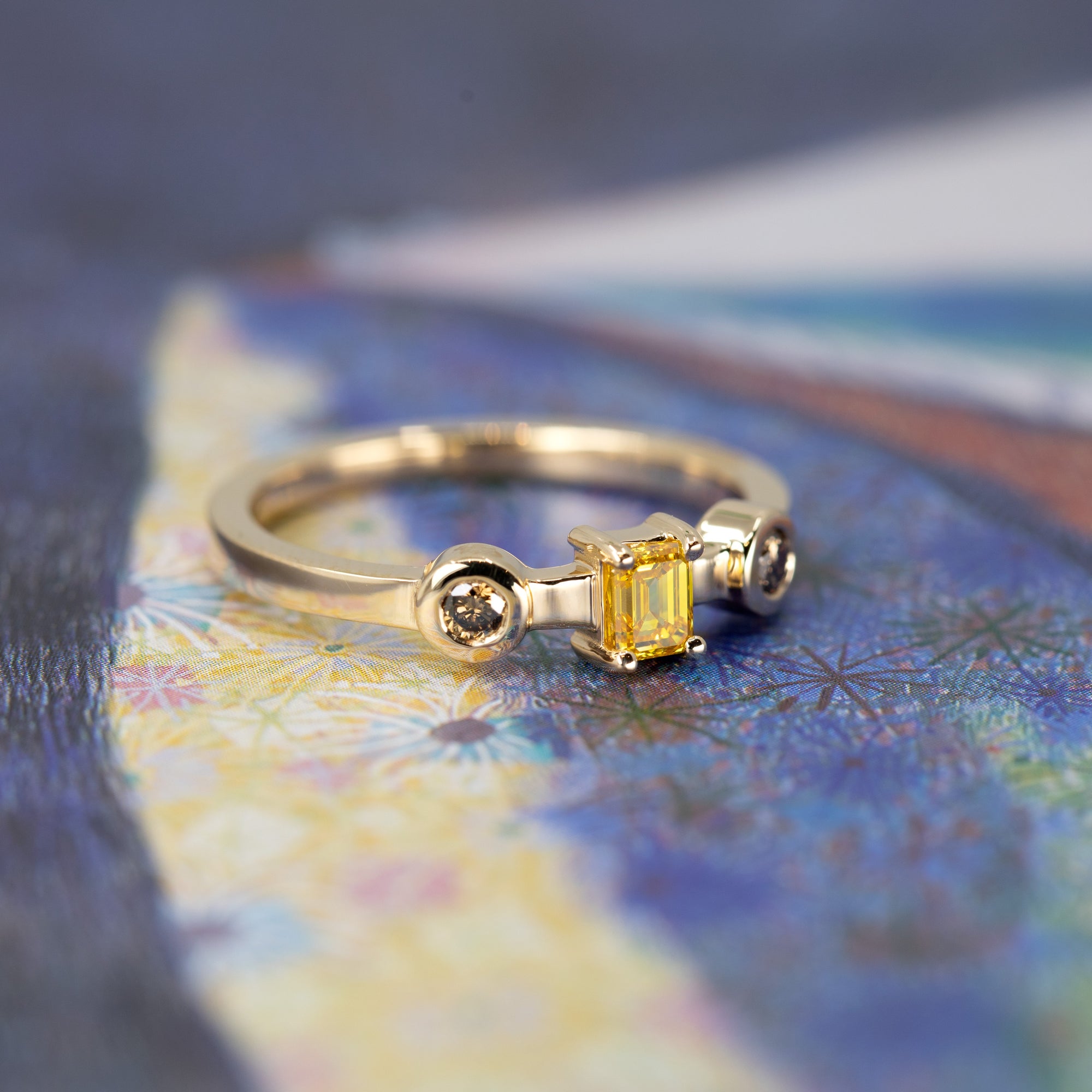 Artistic composition of a yellow gold ring with an emerald-cut yellow diamond and brown side diamonds, showcased on a soft-toned colored background.