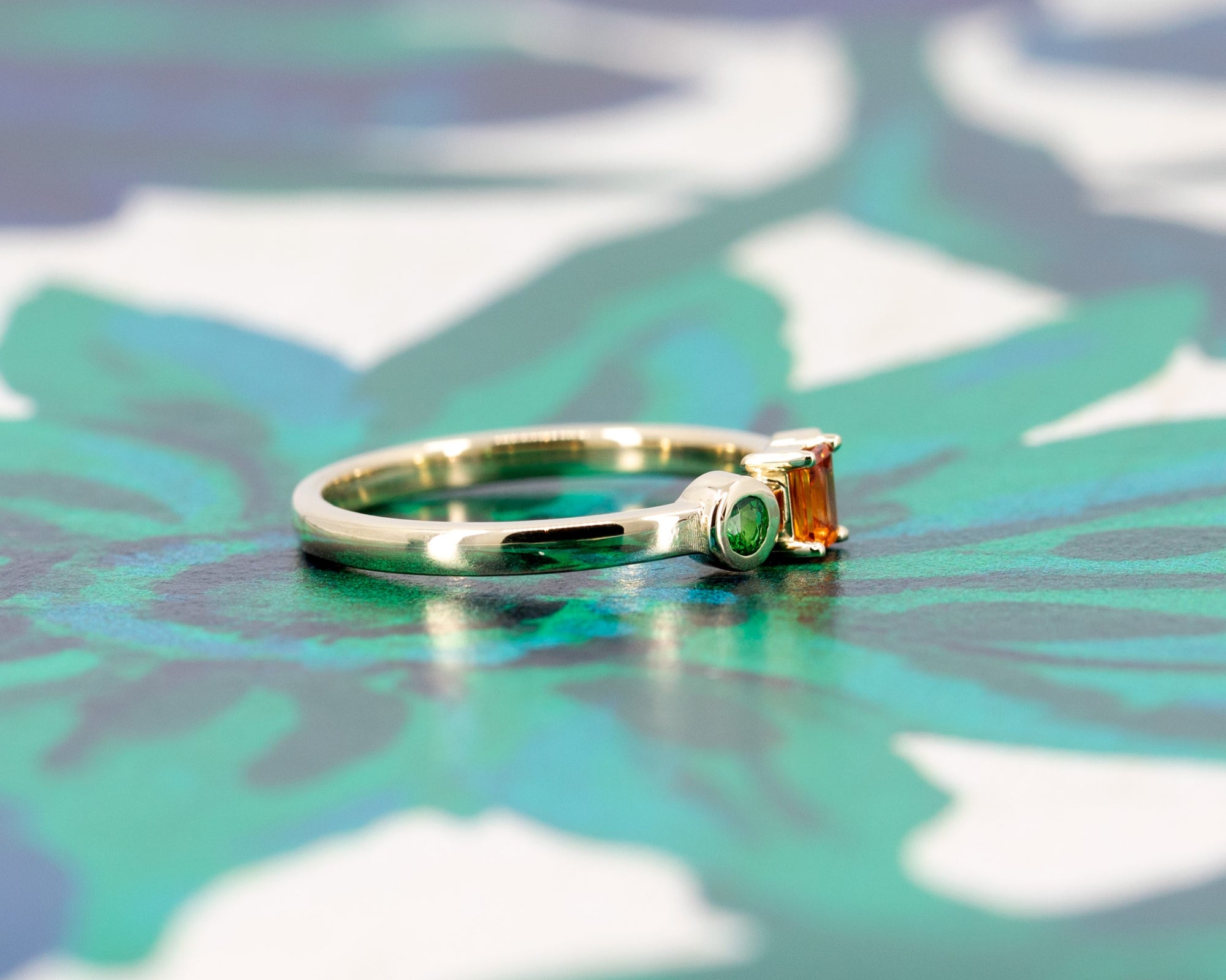 Elegant yellow gold ring with an orange sapphire and green garnet accents, displayed on a soft-toned background. Designed by Bena Jewelry in Montreal.