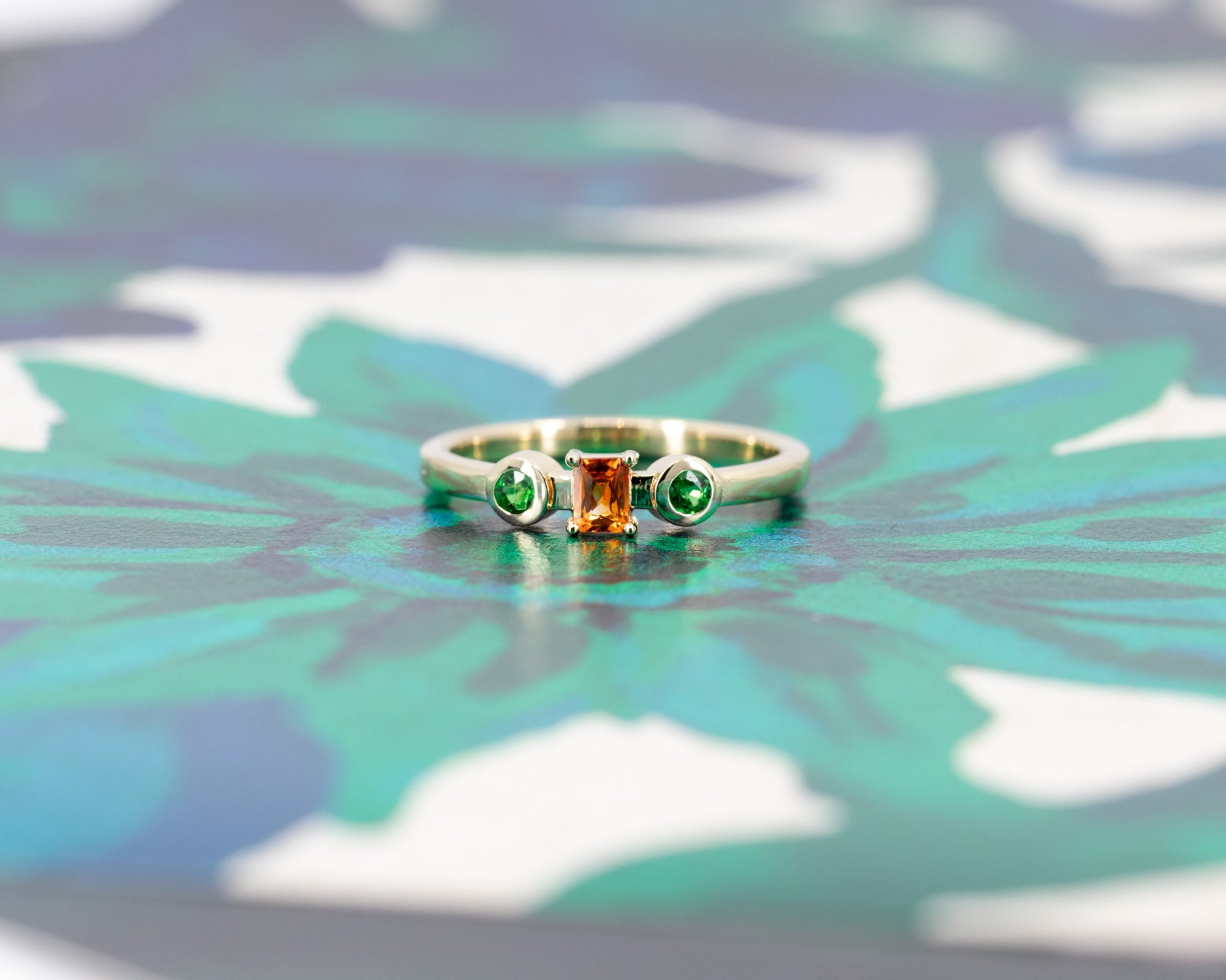 Modern yellow gold ring with vibrant orange and green gemstones, photographed against a warm-colored background. Custom-made by Bena Jewelry in Montreal.
