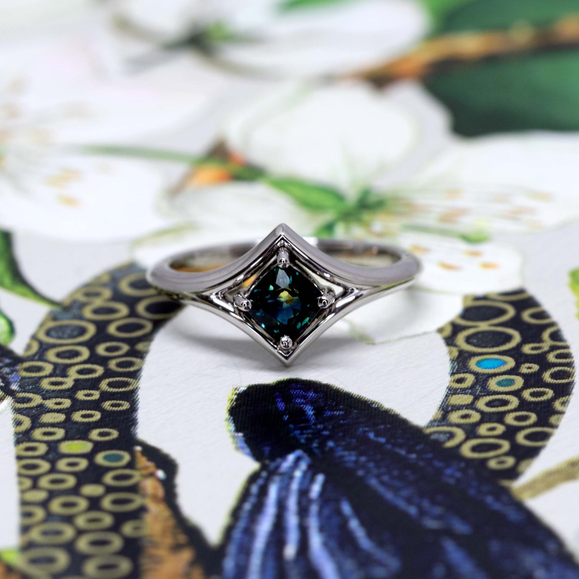Vivid tones surround this striking cushion-cut teal sapphire ring, capturing the elegance and modern aesthetic of Bena Jewelry.