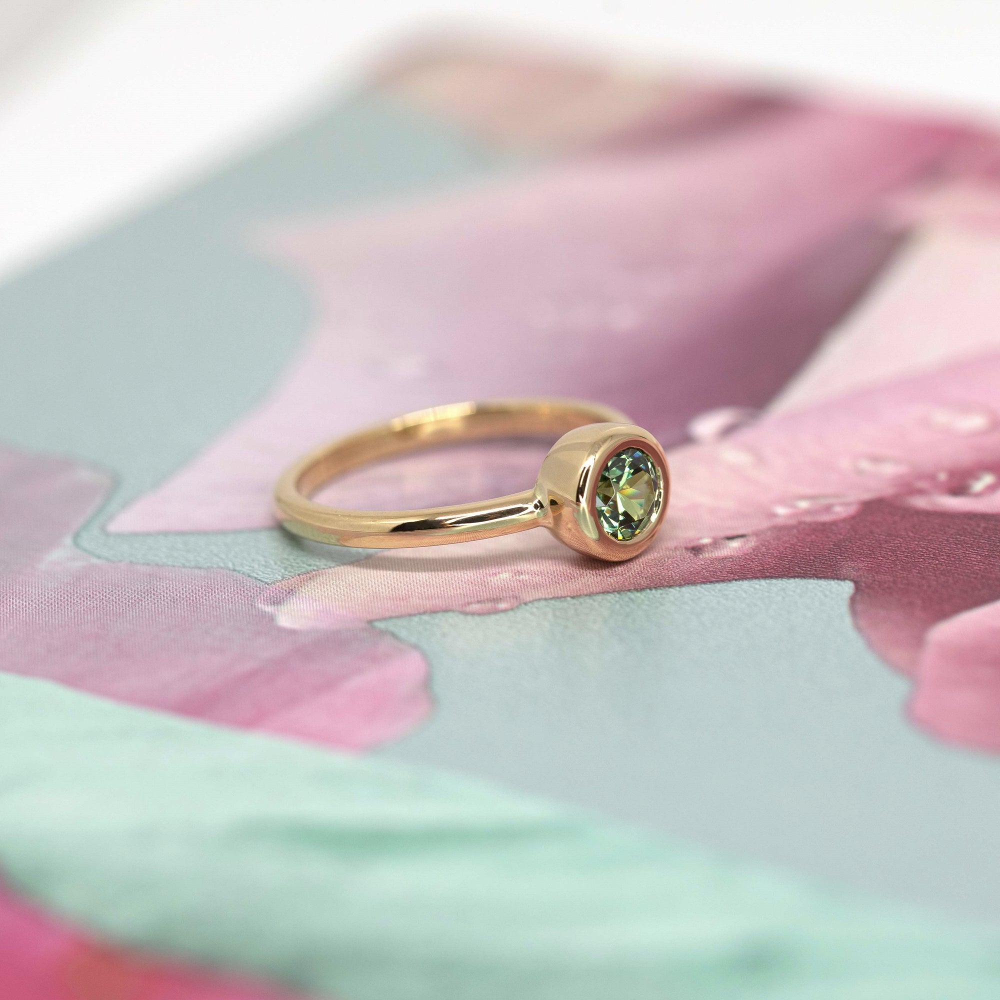 Yellow gold ring with a bezel-set green demantoid garnet by Bena Jewelry, shown on a rose-hued floral background. Minimalist bridal jewelry.