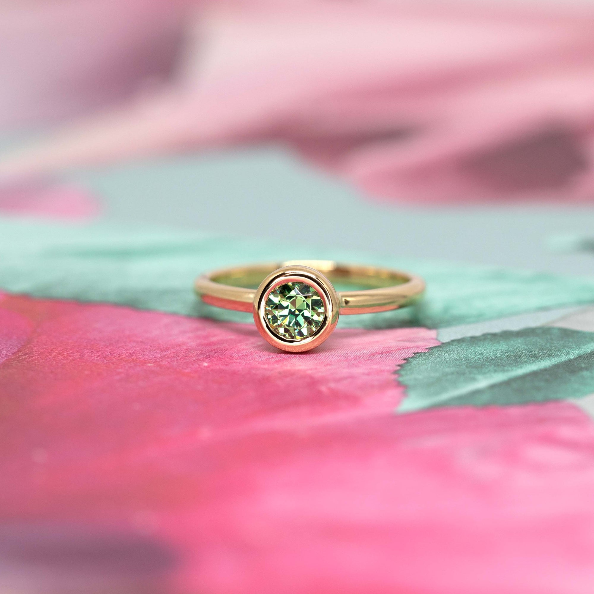 Bena Jewelry’s Bezel Set Demantoide Ring, a minimalist yellow gold ring with a green garnet, displayed on a delicate pink flower-themed background.