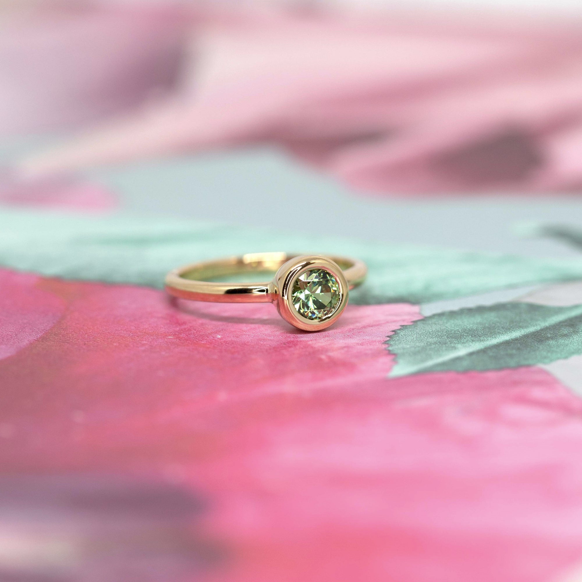 Bezel Set Demantoide Ring by Bena Jewelry, with a vibrant green garnet set in yellow gold, displayed on a soft pink floral background. Handmade in Montreal.