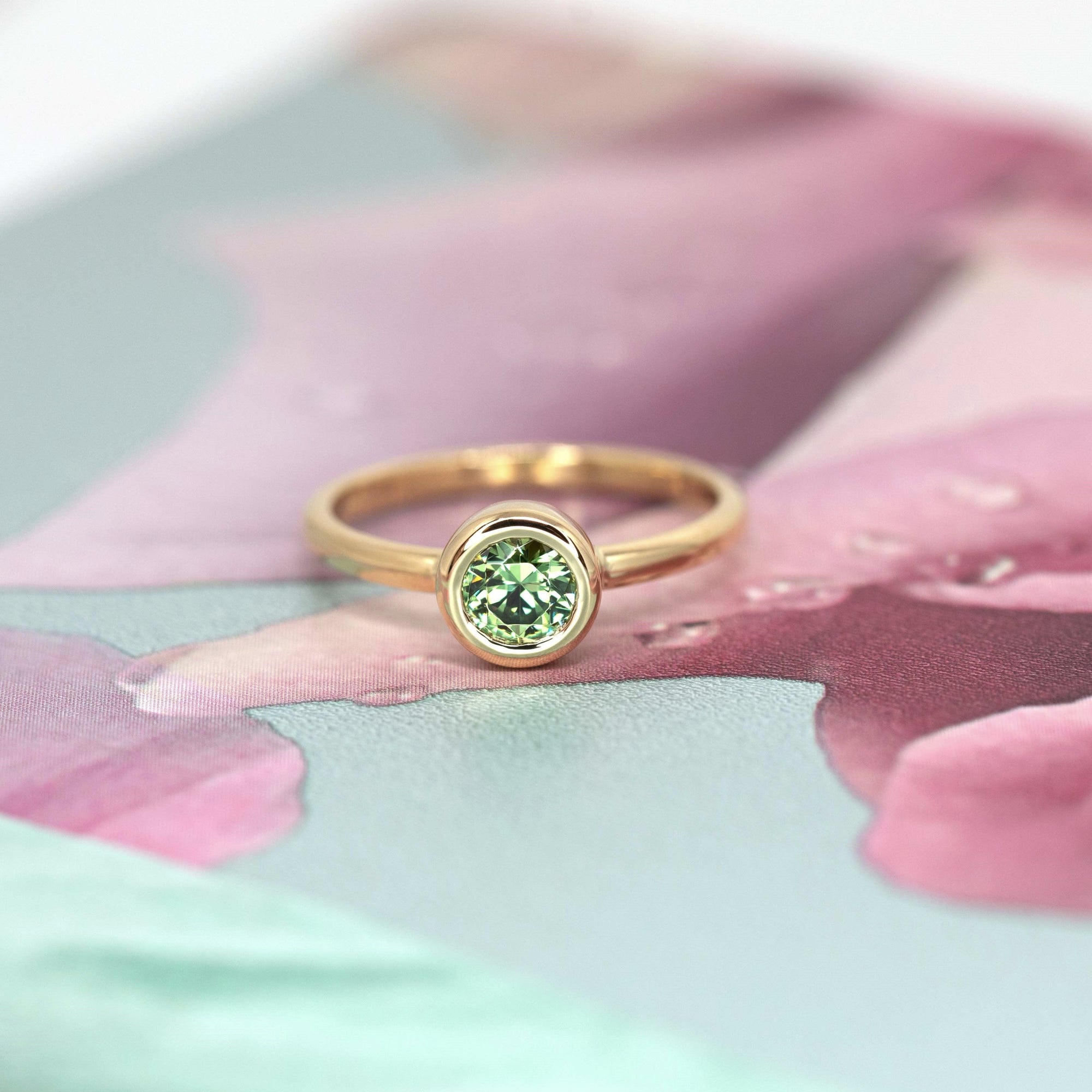 Green demantoid garnet engagement ring by Bena Jewelry, photographed against a romantic pink floral backdrop. Custom-made in Montreal.