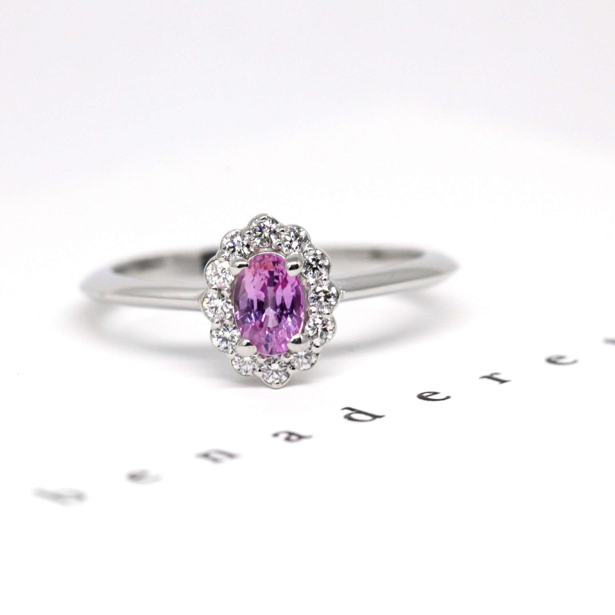 Diamond Halo Pink Sapphire Ring in white gold featuring an oval pink sapphire surrounded by diamonds, photographed on a white background, custom bridal jewelry by Bena Jewelry.