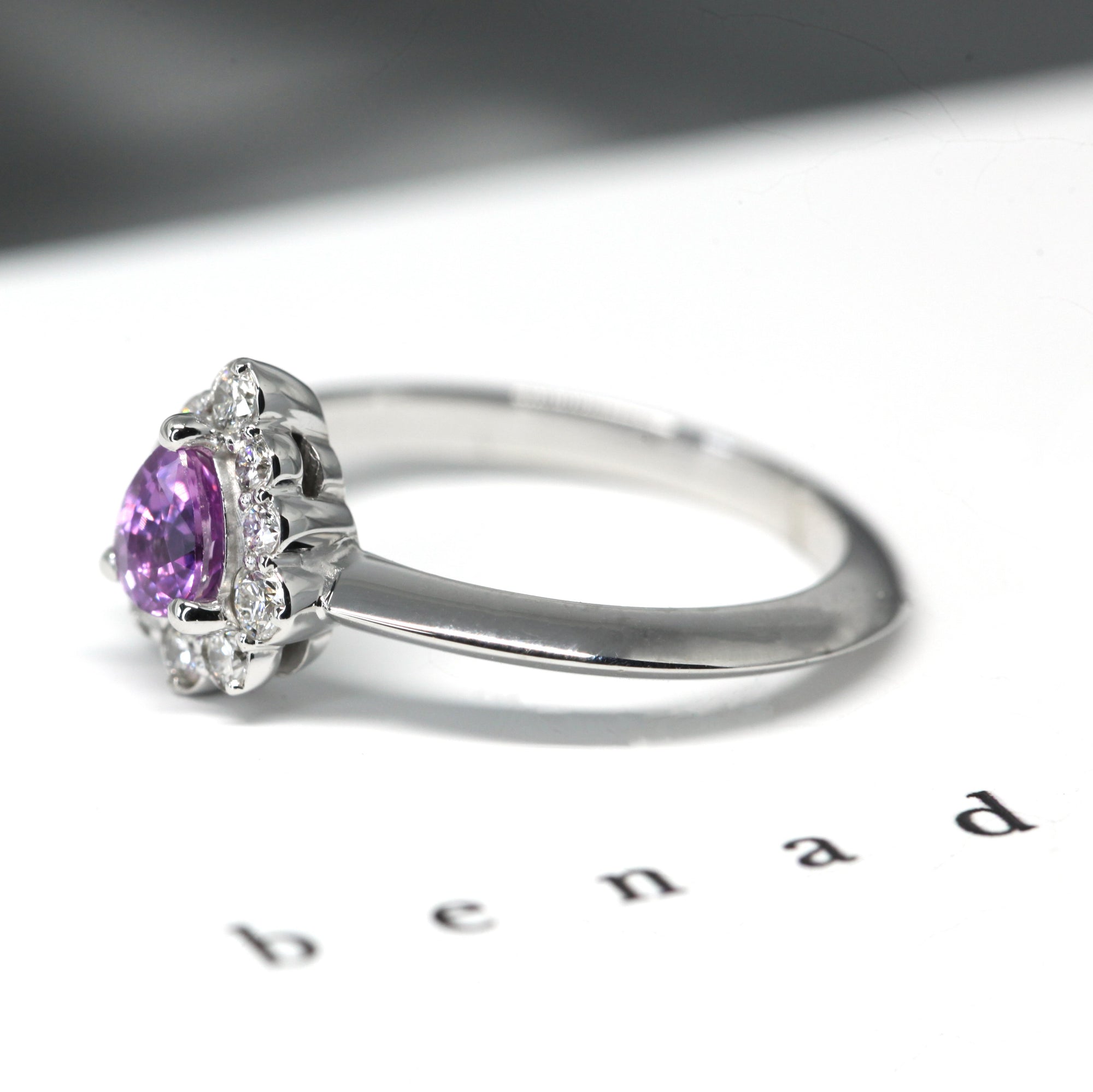 Custom-made bridal ring in white gold, featuring a mesmerizing pink-violet sapphire and diamond halo. Crafted by Bena Jewelry.