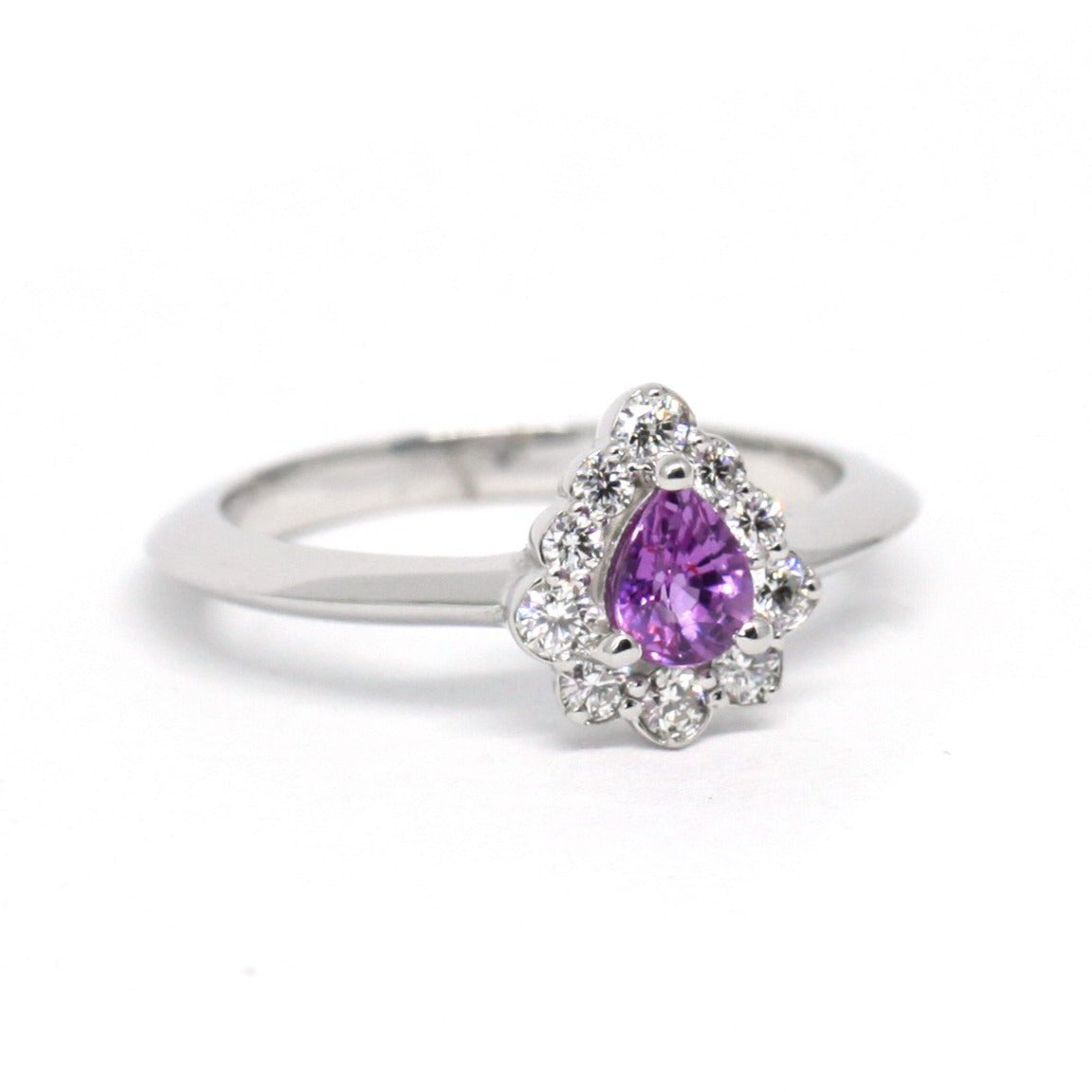 Elegant sapphire and diamond halo ring crafted in white gold, showcasing a custom pear-cut pink-violet sapphire centerpiece. Designed by Bena Jewelry, an independent Canadian jewelry designer in Montreal.