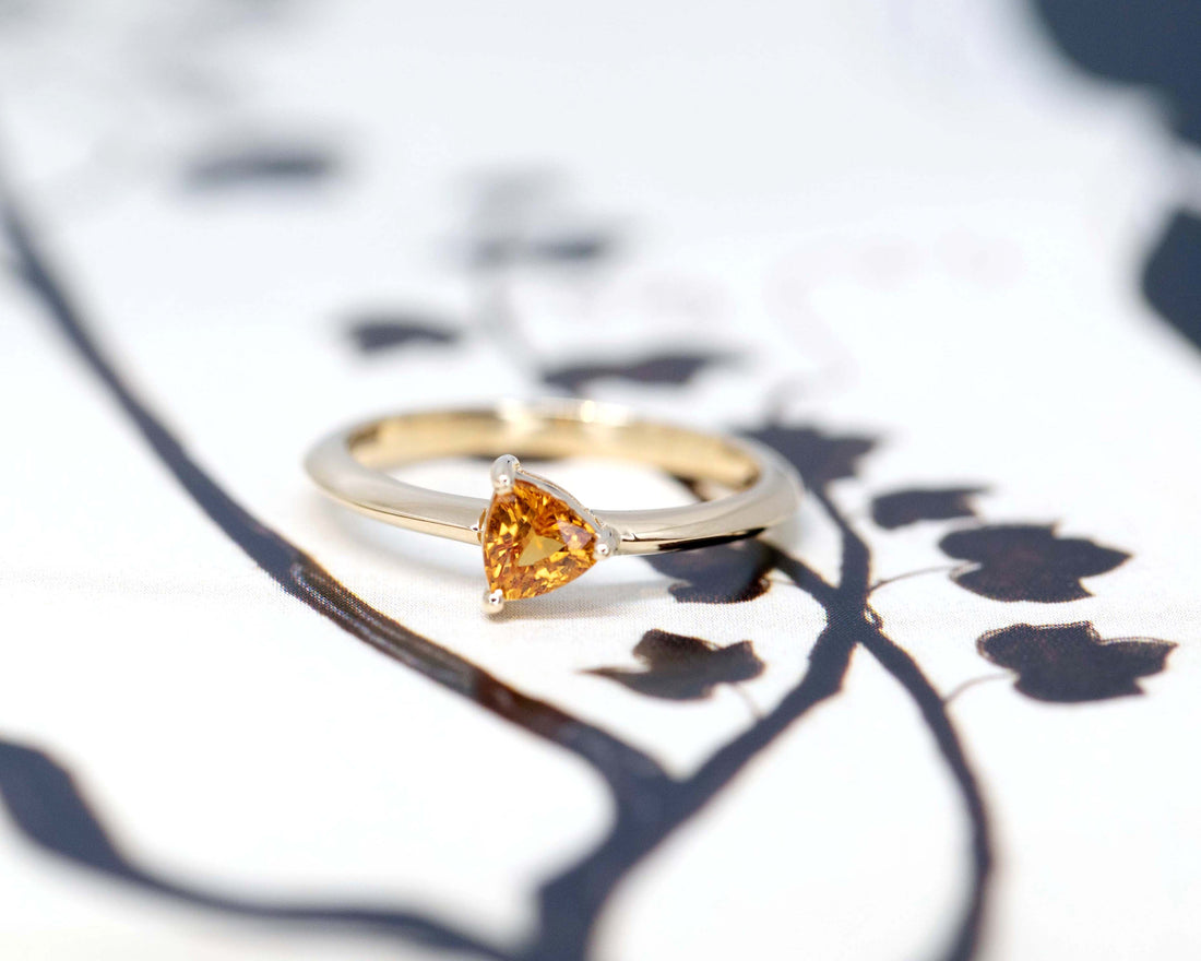 front view of a custom made minimalist bridal ring with a orange trillion shape sapphire made in montreal by bena jewelry designer on a black and white background